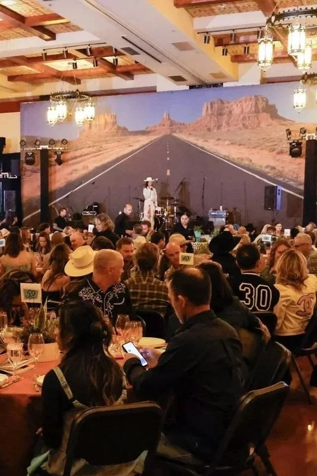 A large indoor event with many people seated at tables, and a stage with a desert road backdrop and rock formations. There are musical instruments and speakers on the stage.