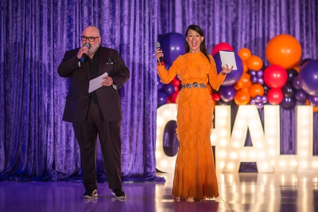Man and Yve Rojas on stage with colorful balloons and large illuminated letters in the background, Yve in an orange dress holding a microphone and smiling, man in a black suit holding a paper and microphone.