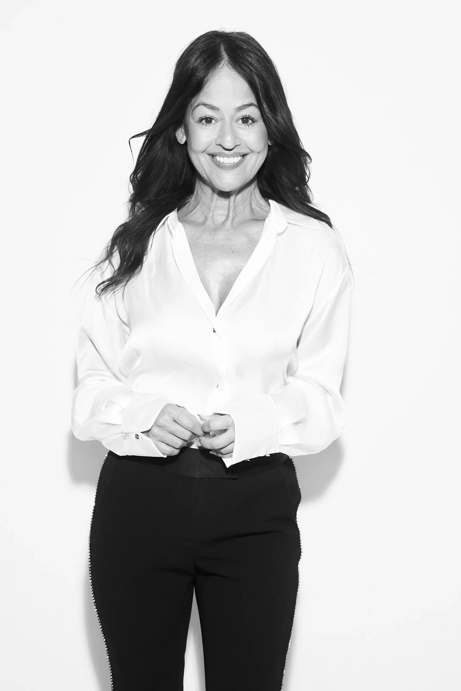 Black and white photo of a smiling Yve Rojas with long dark hair, wearing a white blouse and black pants, standing against a plain background.