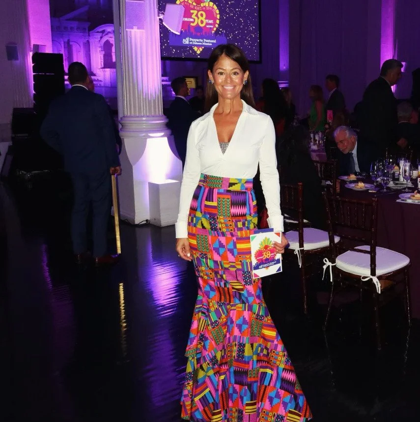 Yve Rojas in a white blouse and a colorful, patterned maxi skirt stands in a decorated event hall with purple lighting, holding a program or brochure, smiling at the camera.