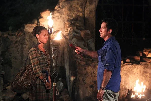 Yve Rojas with dark hair in a bun wearing a plaid shirt and carrying a large leopard print bag stands outdoors at night. A man in a blue shirt is holding a red object towards her. 