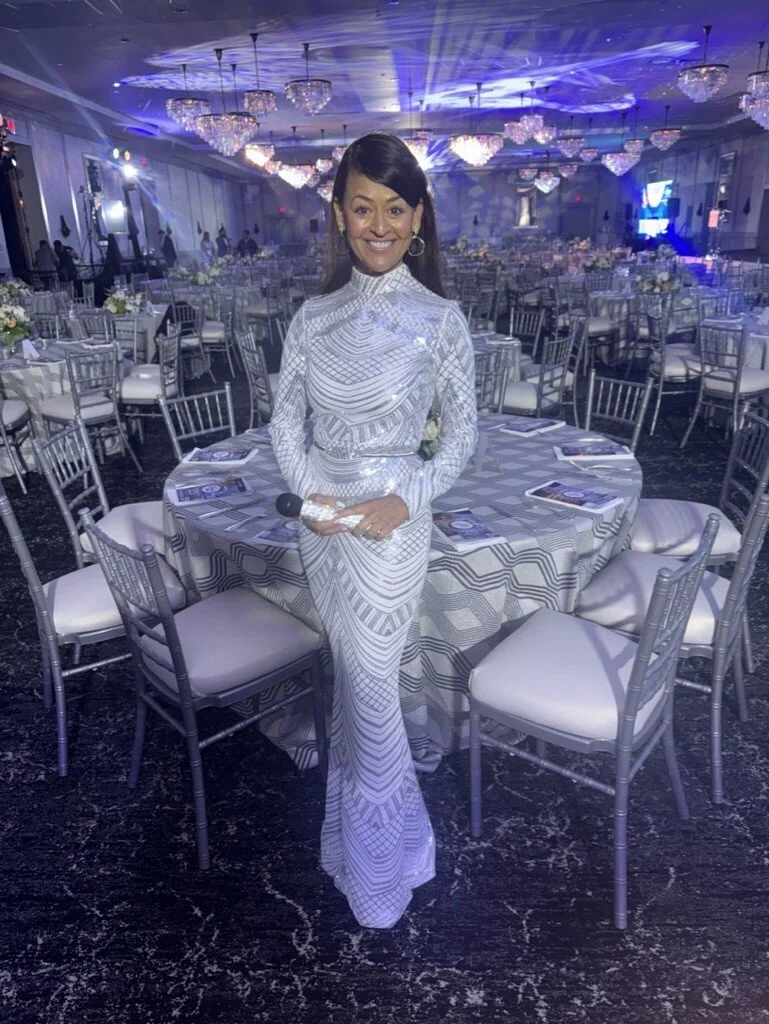 Woman in a shiny, silver, patterned dress standing in a decorated banquet hall with round tables, calligraphed menus, and chandeliers, holding a microphone and smiling.