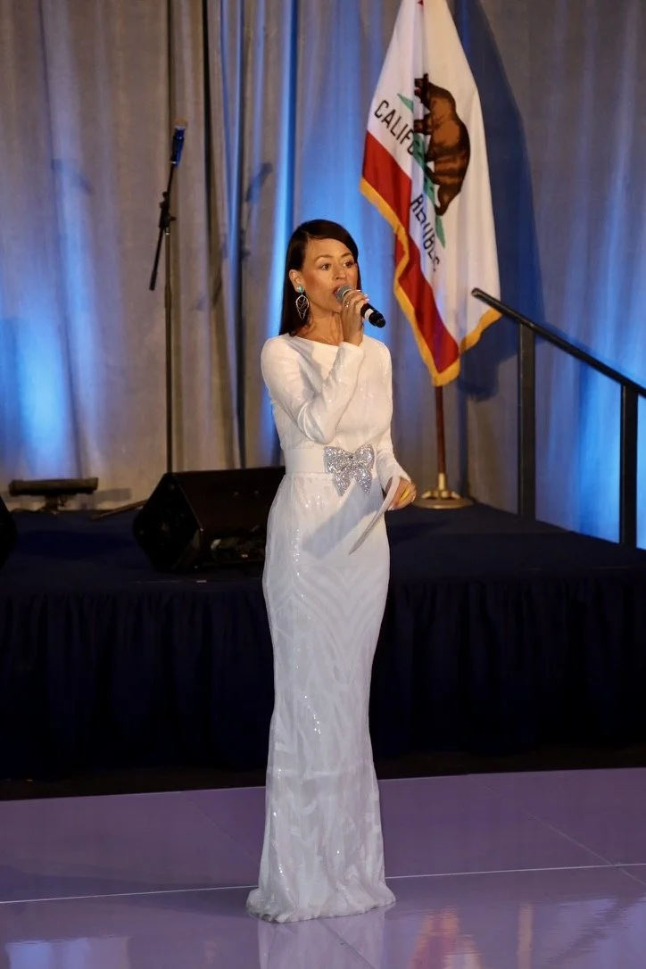 Yve Rojas in a white gown with a silver bow is standing on a stage, speaking into a microphone. Behind her is a California state flag and grey curtains, with blue lighting.