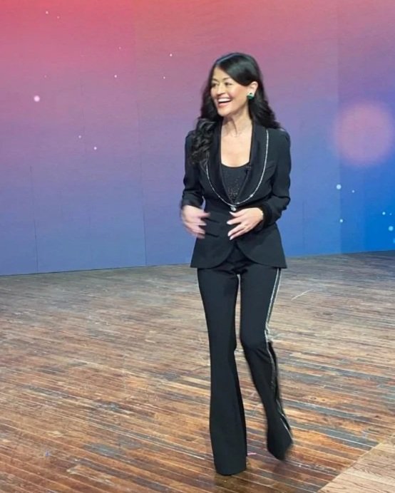Yve Rojas in a black suit with stripe pants, smiling and walking on a wooden stage with a pink and purple background.
