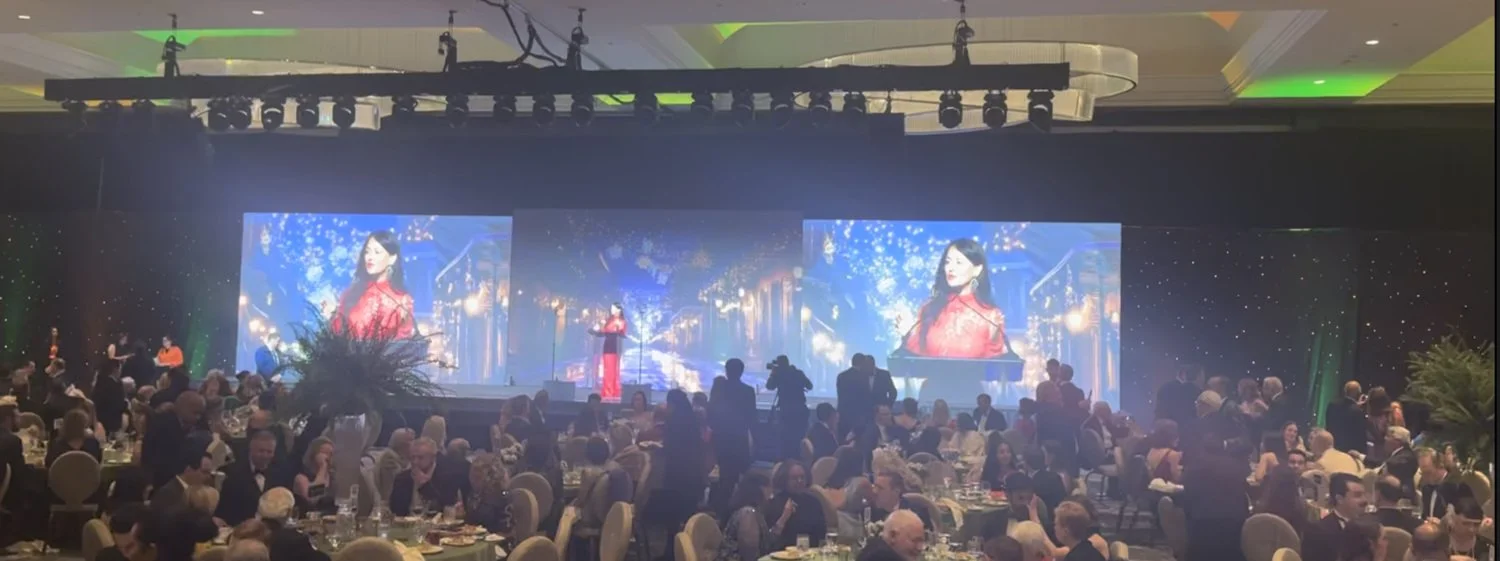Large ballroom filled with round tables and many people seated, with a stage at the front displaying two large screens showing Yve Rojas in a red outfit, and a Yve standing on stage speaking into a microphone.