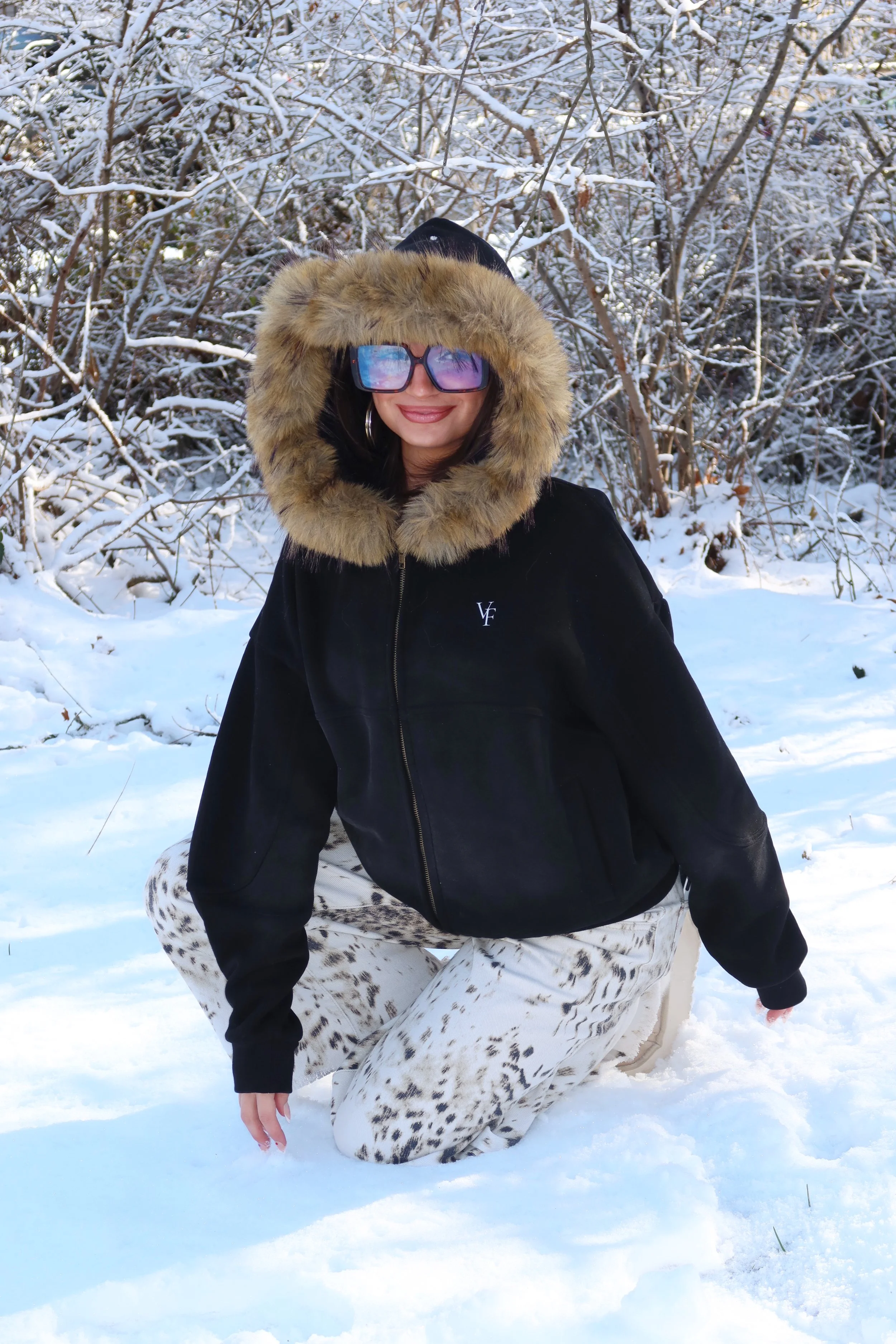A woman dressed in winter clothing, including a black jacket with a fur-lined hood, leopard print pants, and large blue sunglasses, kneeling on snow in a snowy outdoor setting with snow-covered trees in the background.
