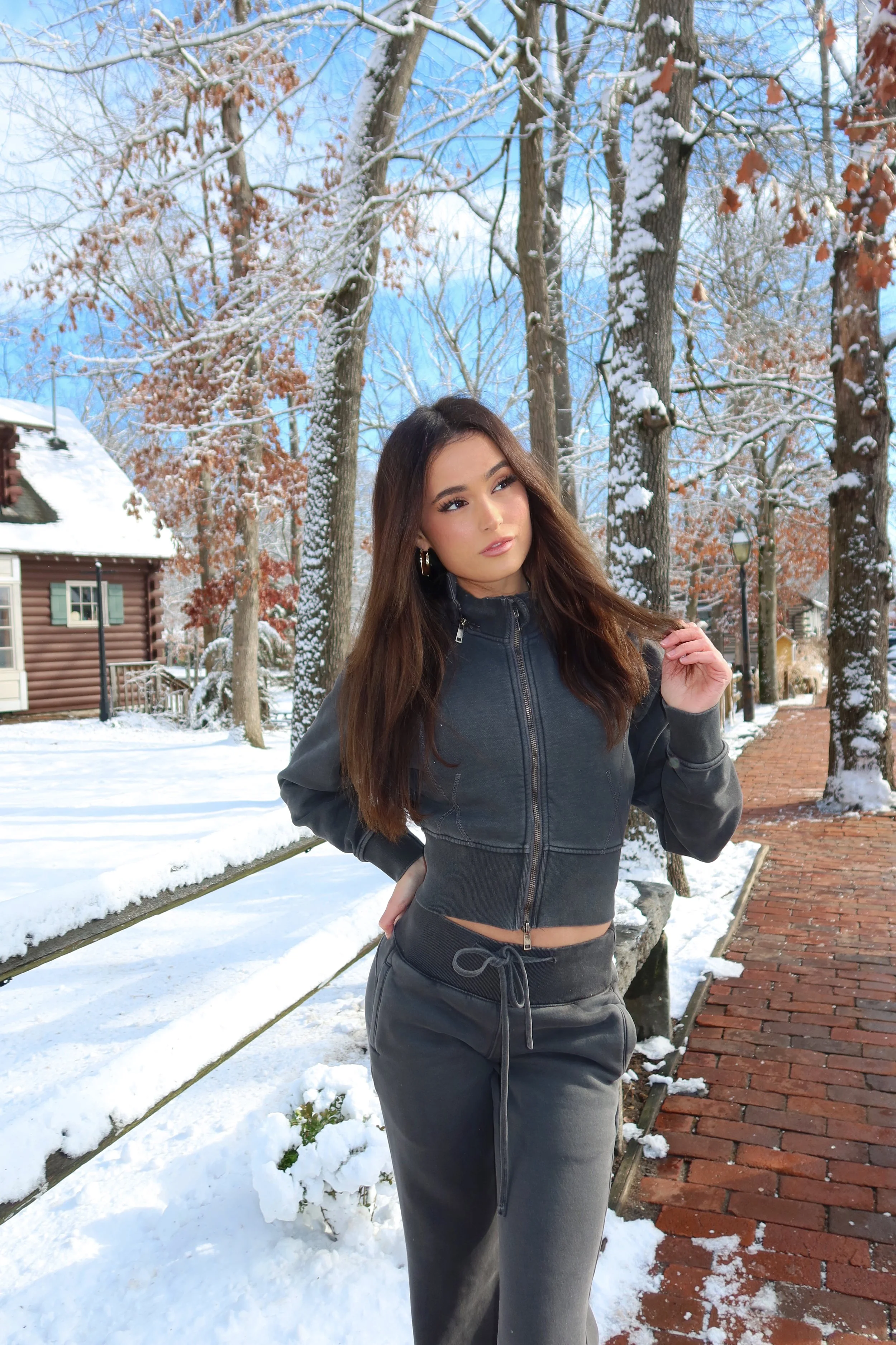 Young woman in black zip-up hoodie and sweatpants standing on snowy brick sidewalk in a winter neighborhood with snow-covered trees and houses.