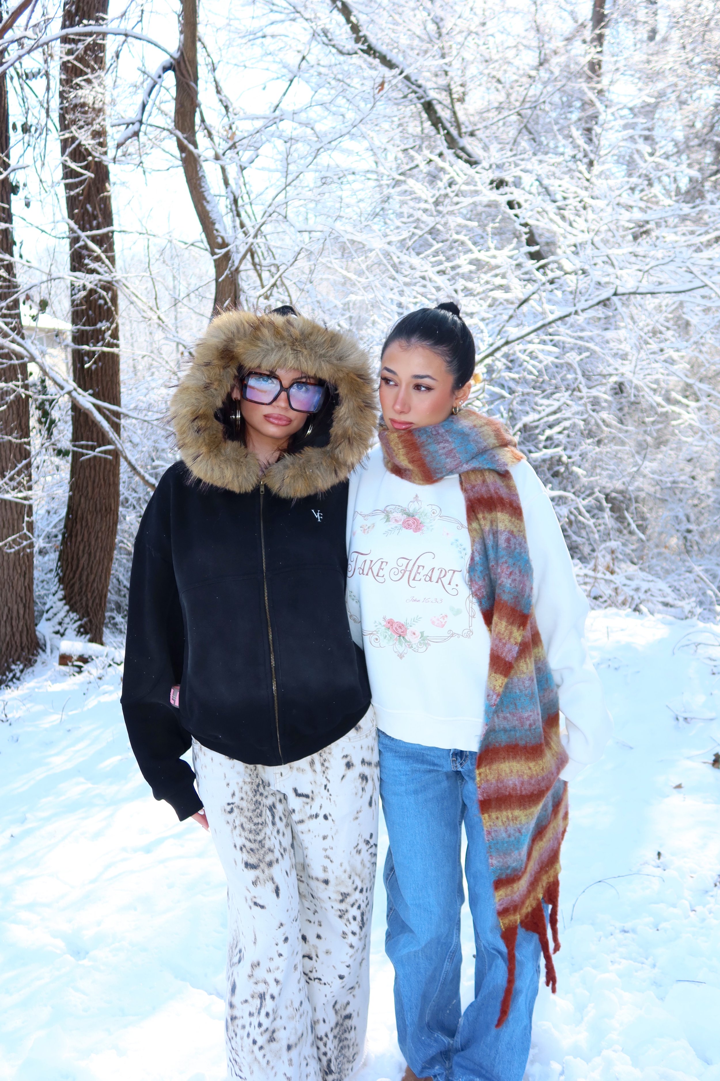 Two women standing outdoors in a snowy forest, dressed in winter clothing. One woman is wearing a black jacket with a fur-lined hood and large glasses, and the other is wearing a white sweatshirt with floral design, a colorful scarf, and jeans.