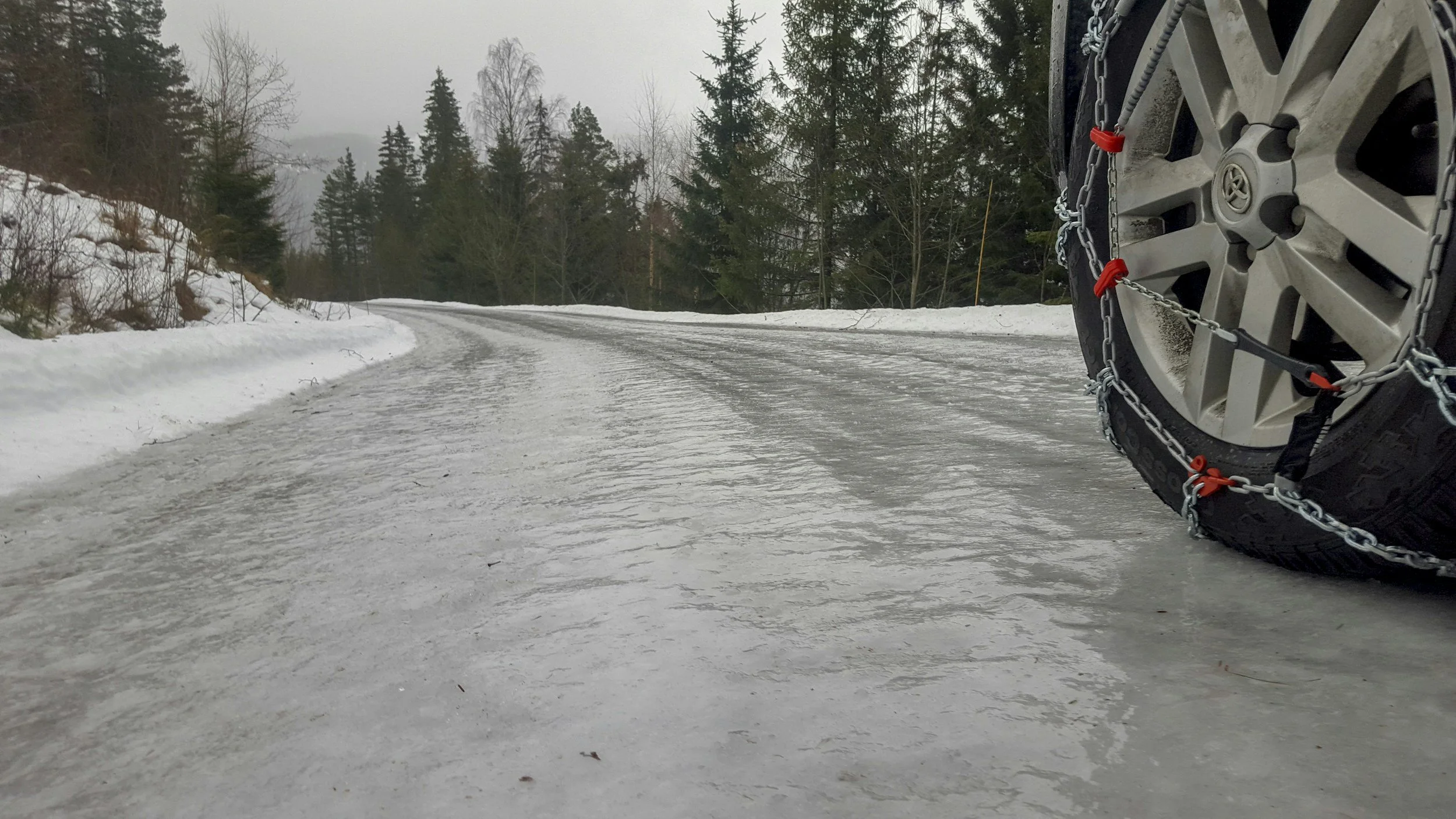 Close-up of a car tire with metal chains, driving on an icy mountain road with snow and evergreen trees in the background.