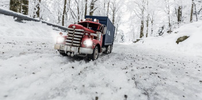 A red semi-truck driving on a snowy road in a winter forest.