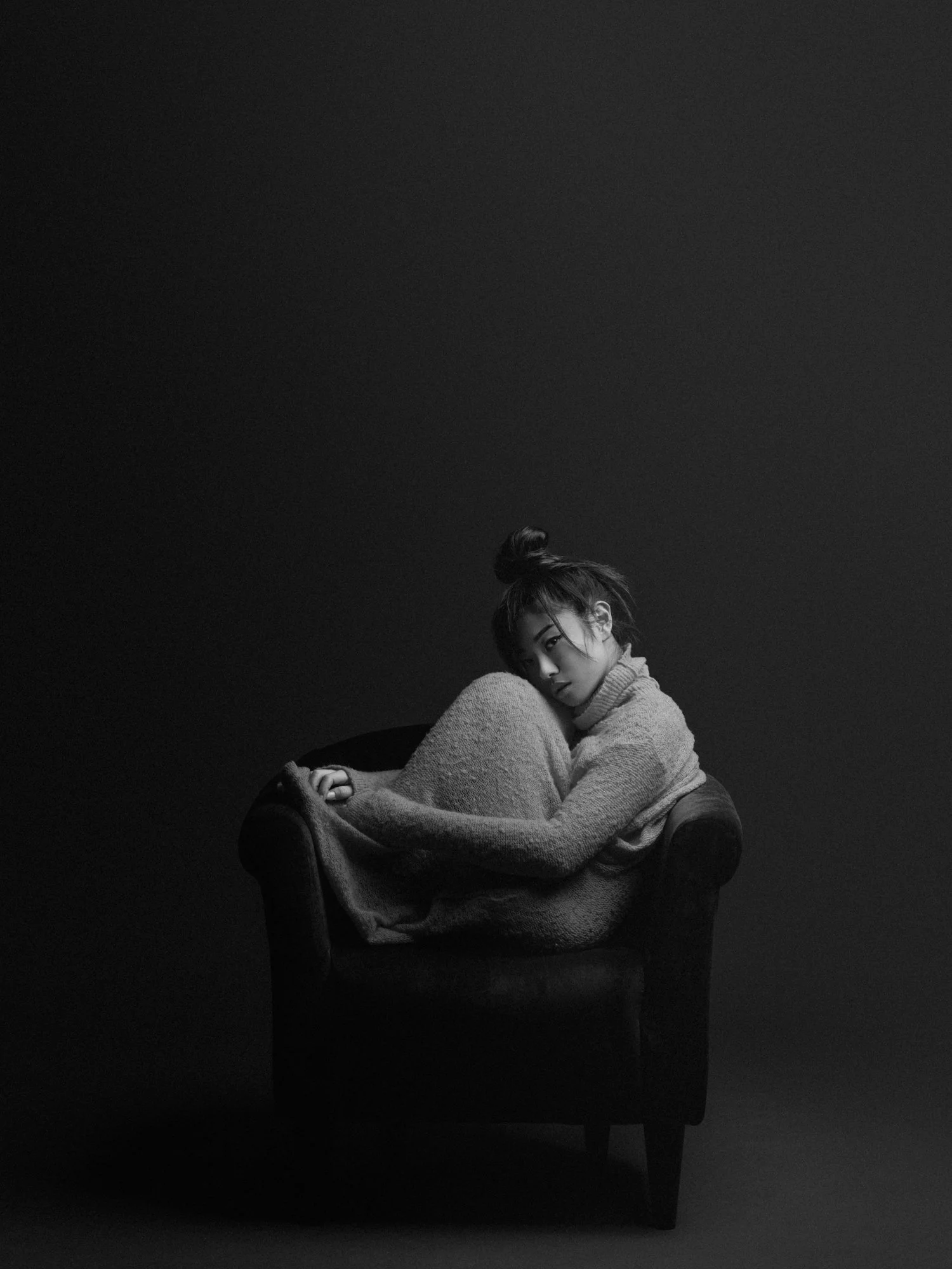 Black and white photo of a woman sitting on a chair in a dark room, with her knee drawn up and her head resting on her knee, looking at the camera.
