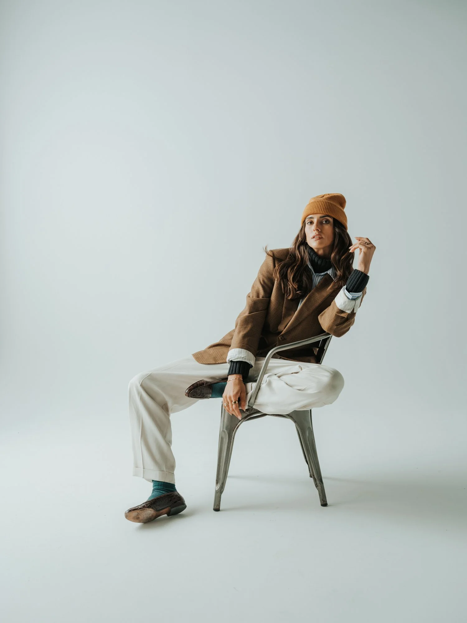 A woman with long dark hair, wearing a brown cap, brown blazer, black turtleneck, white pants, and brown shoes, sitting on a metal chair against a plain light gray background.
