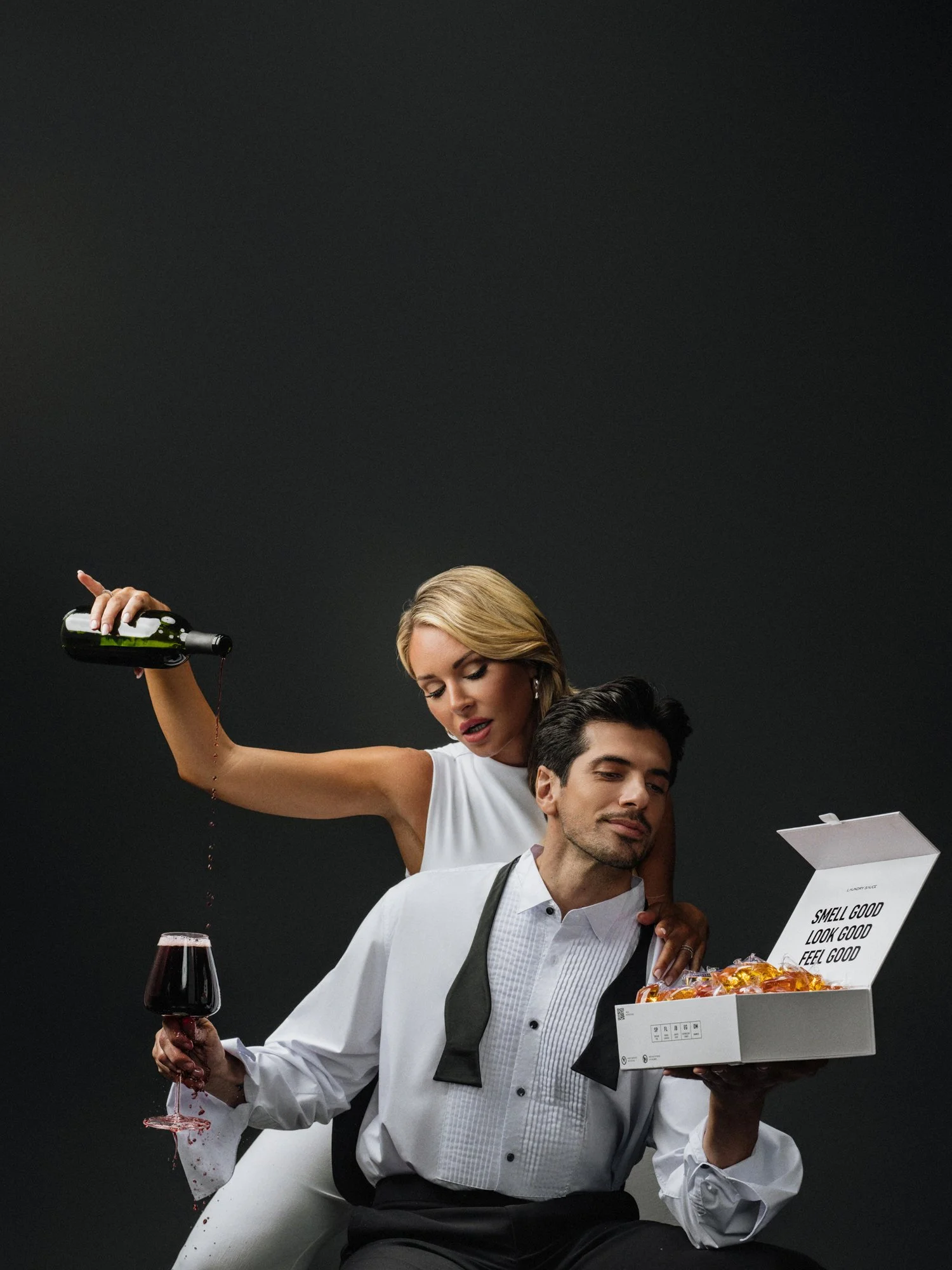 A woman with blonde hair pouring red wine over a man's head while they sit in a white bathtub. The man is holding a glass of red wine. The woman is leaning over him, and they are both dressed formally, with the man in a tuxedo and the woman in a whit