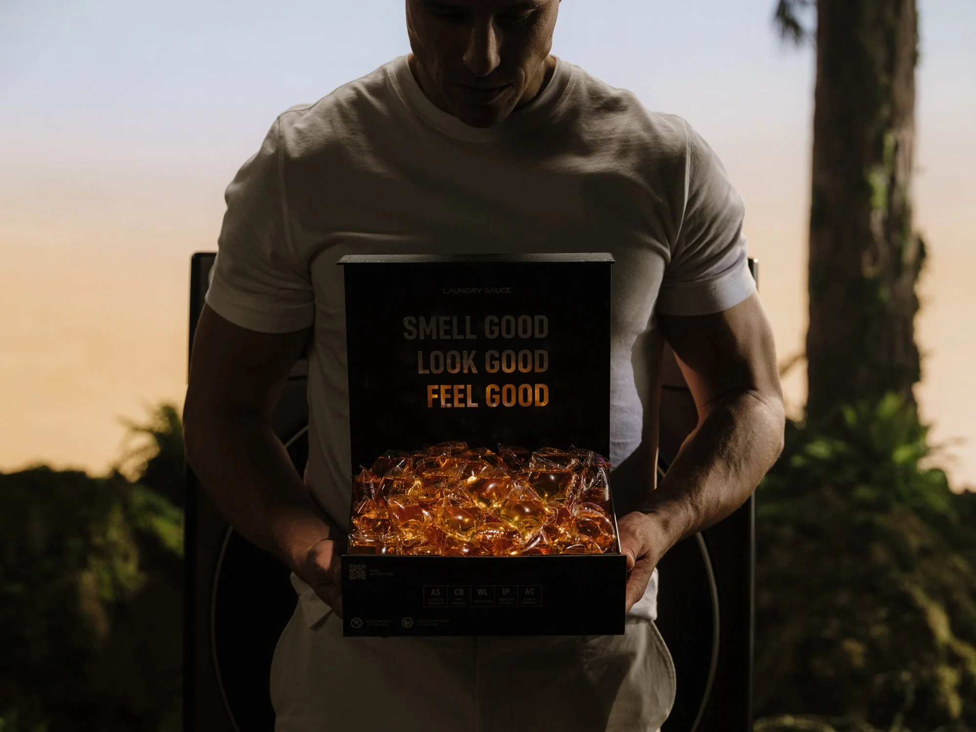 A man in a white t-shirt stands outdoors at sunset, holding an open box filled with glowing amber-colored items, with a sunset sky and trees in the background.
