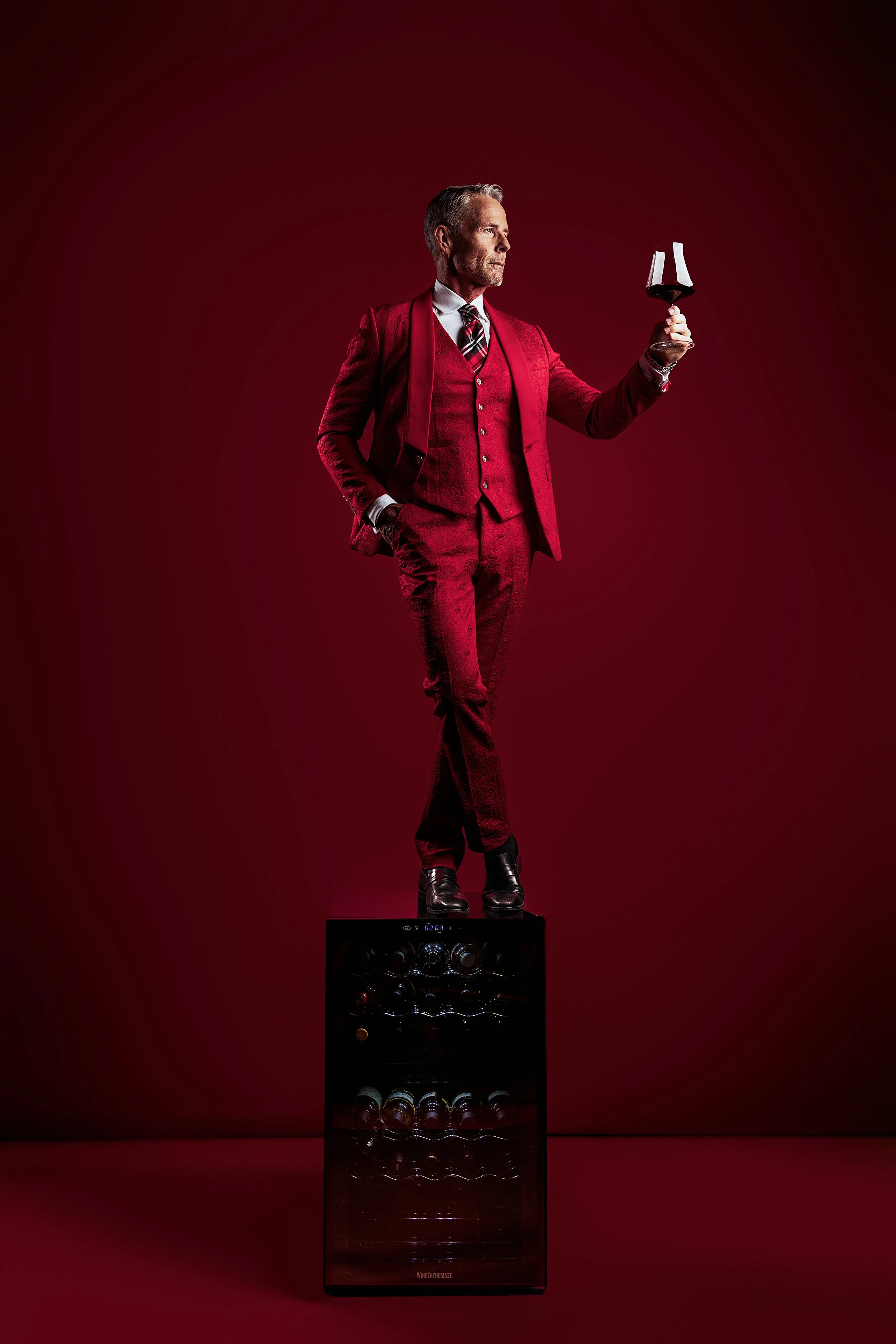 A man dressed in a red suit, white shirt, and plaid tie standing on a wine refrigerator while holding a glass of red wine. The background is deep red, matching his suit.
