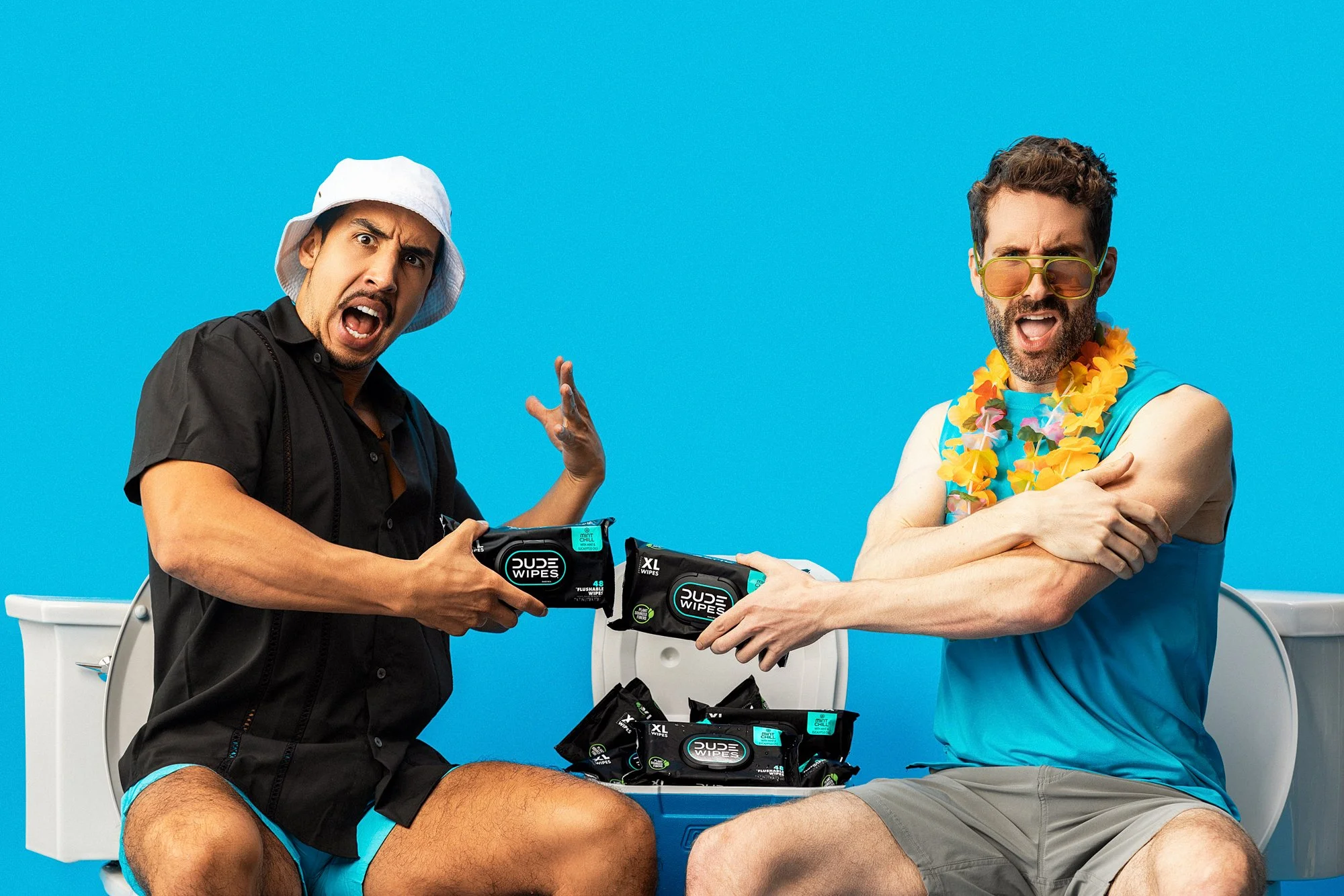 Two men in swimwear sitting on washing machines against a blue background; one wearing a white hat and black shirt, the other with sunglasses and yellow flower lei; they are exchanging packs of DUDE wipes.