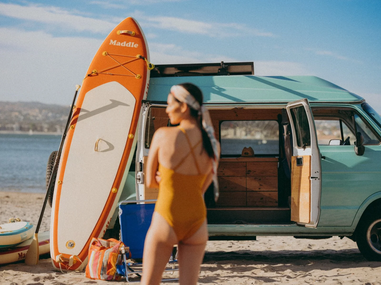 A woman in a yellow swimsuit standing on a beach with a vintage teal van in the background. The van's sliding door is open, revealing wooden cabinetry inside. A paddleboard with orange and white colors is propped against the van, and various beach ge