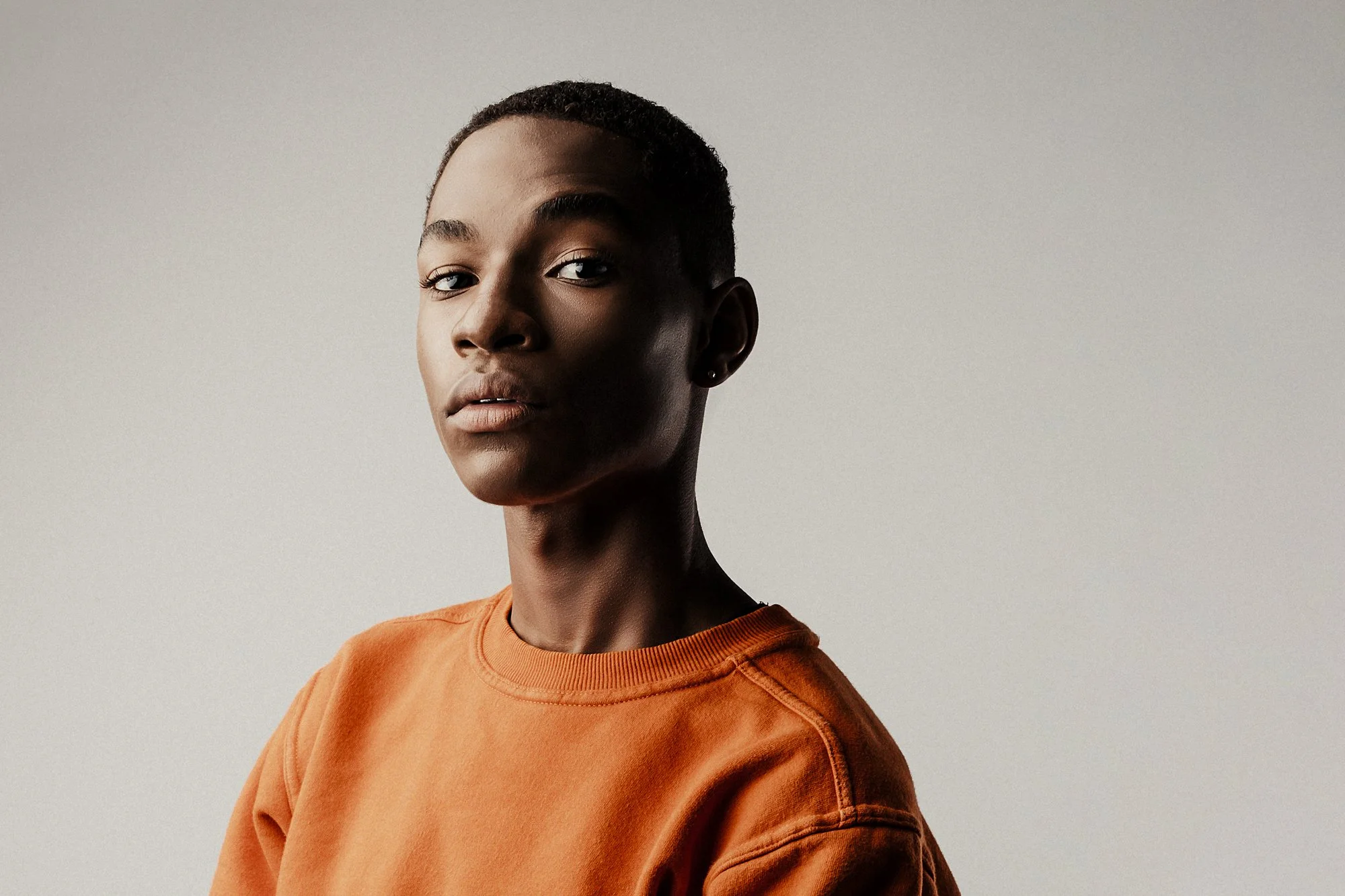 Portrait of a young Black woman with short hair, wearing an orange sweatshirt, against a plain light gray background.