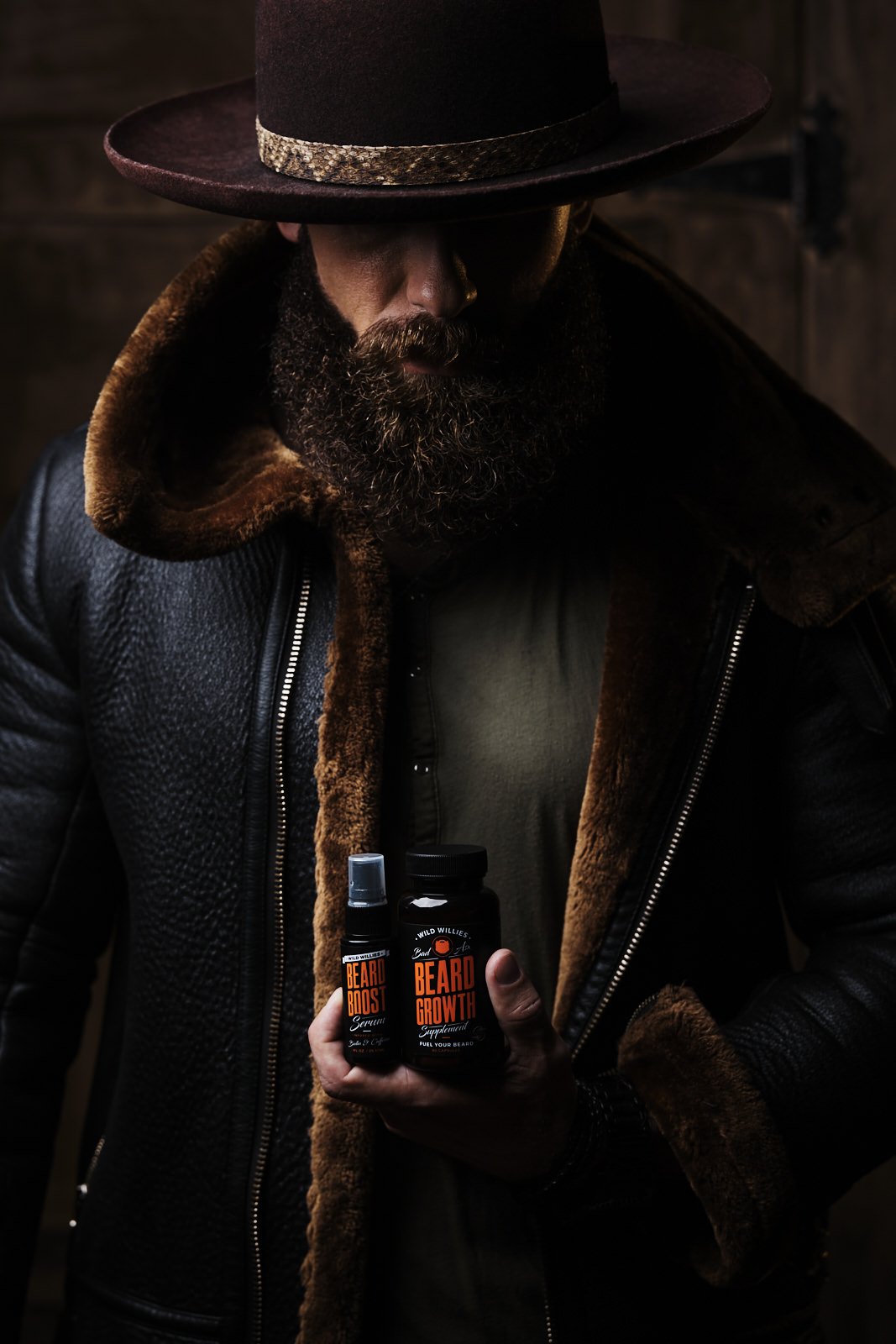 Man with a full beard wearing a wide-brimmed hat and a black leather jacket with brown fur collar, holding two bottles of beard care supplements, looking down.