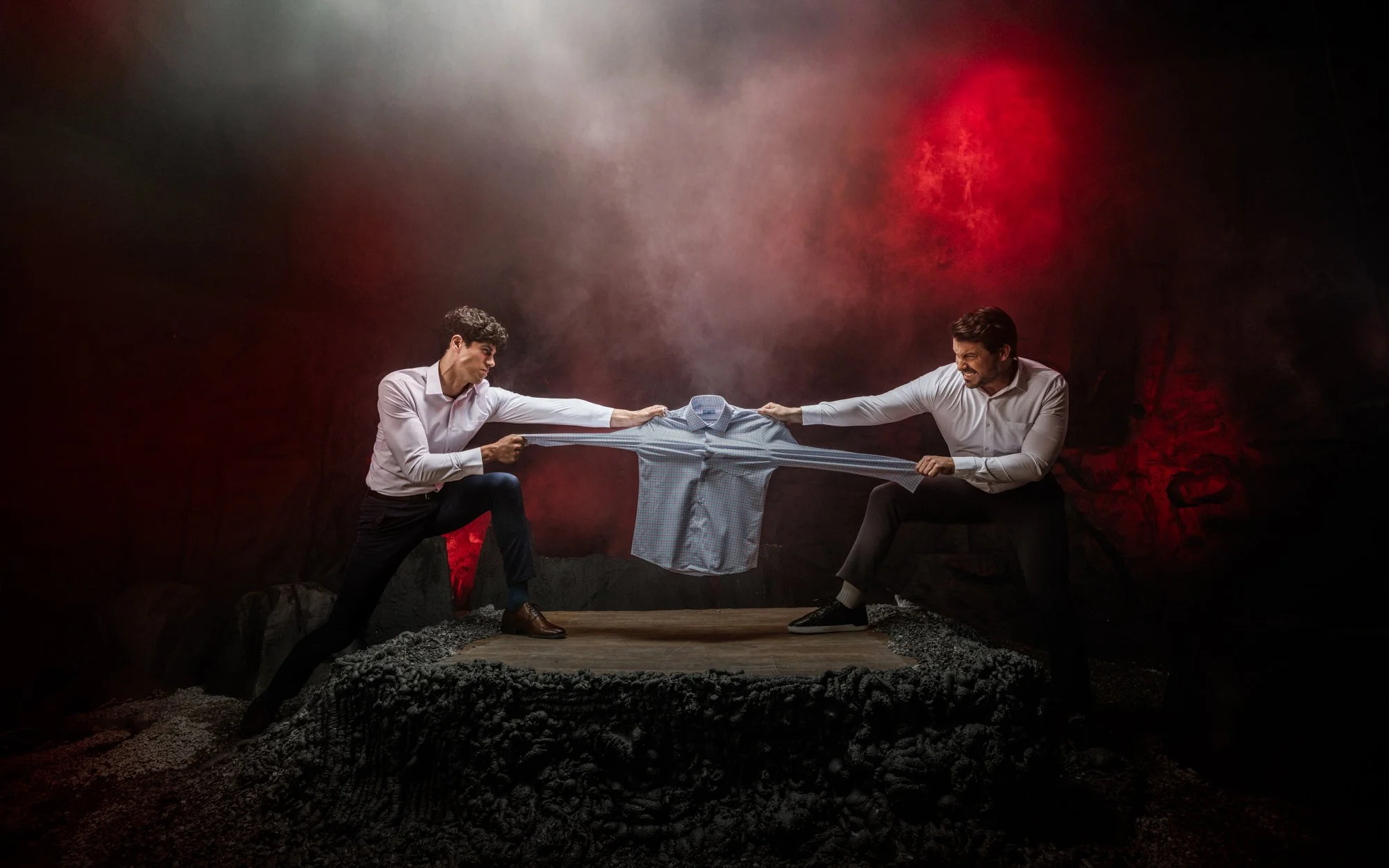Two men in white shirts and dark pants tugging on opposite sides of a shirt, pulling it across their outstretched arms in a staged tug-of-war. They are in a dark, rocky setting with red lighting in the background.