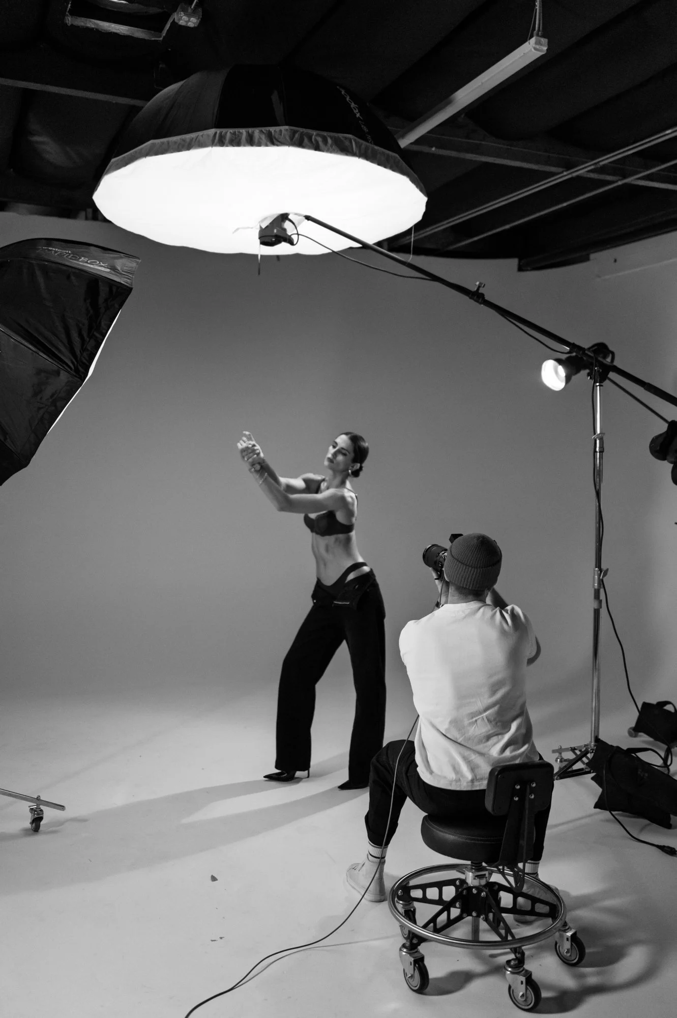 Black and white photo of a photo shoot in progress. A woman is posing in front of a plain background, wearing stylish clothes and high heels. The photographer, dressed in a white hoodie and striped socks, is seated while taking her picture. Large studio umbrellas and lighting equipment surround them, with one umbrella illuminated from above.