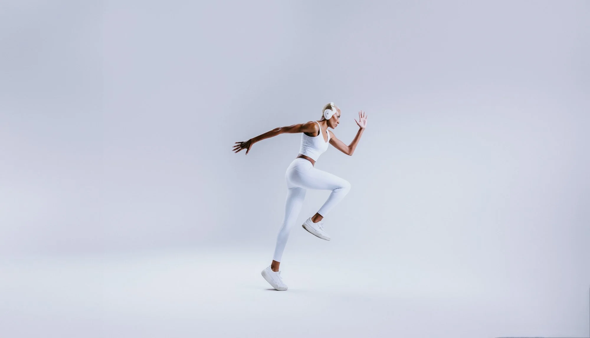 A woman dressed in white activewear, wearing headphones, running or jumping in a minimalistic setting with a plain light gray background.