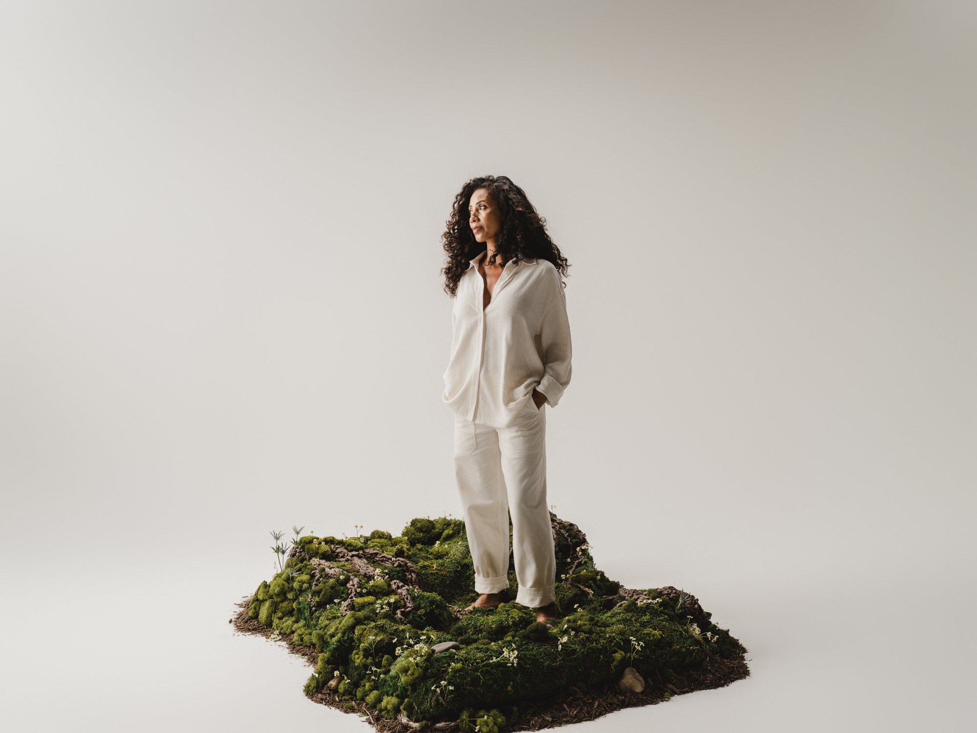 A woman with curly hair wearing a beige shirt and pants standing on a small patch of greenery and rocks, with a neutral background.