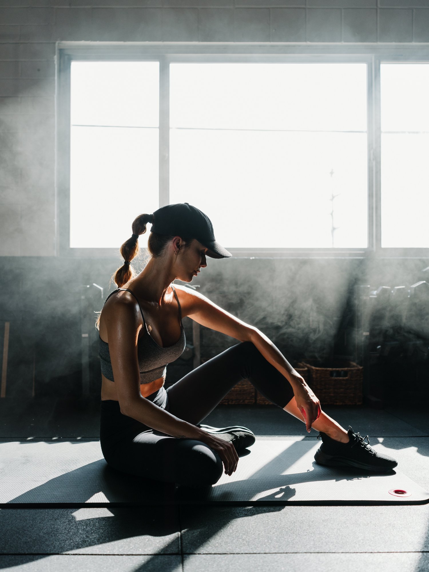 A woman in workout clothes and a cap sitting on a yoga mat in a gym, stretching her leg while facing a large window.