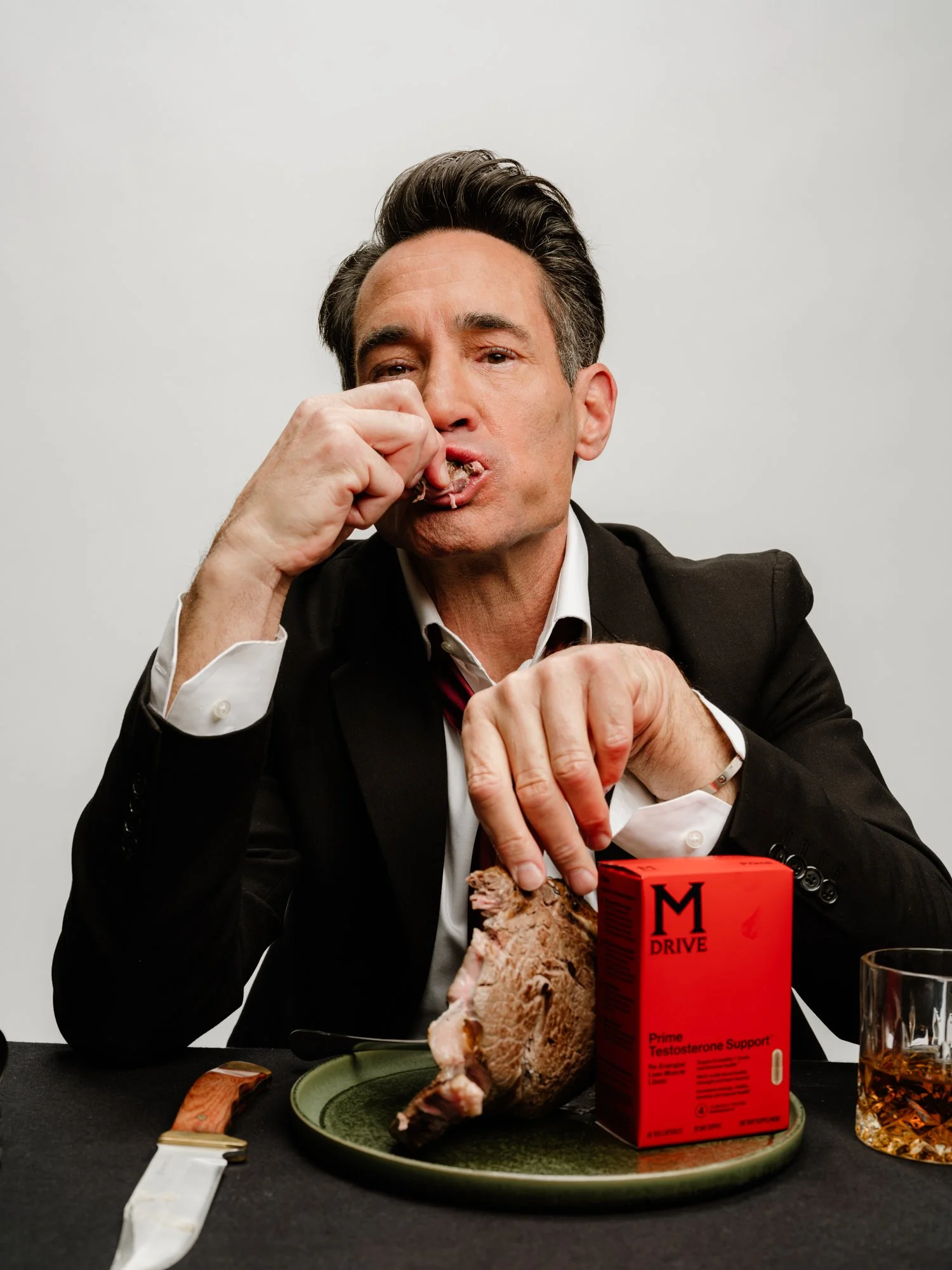 Man in a black suit and white shirt sitting at a table with a large cooked turkey on a green tray, a red box labeled 'Prime Testosterone Support', a glass of whiskey, a knife, and a spoon. The man is eating the turkey with his fingers, appearing to b