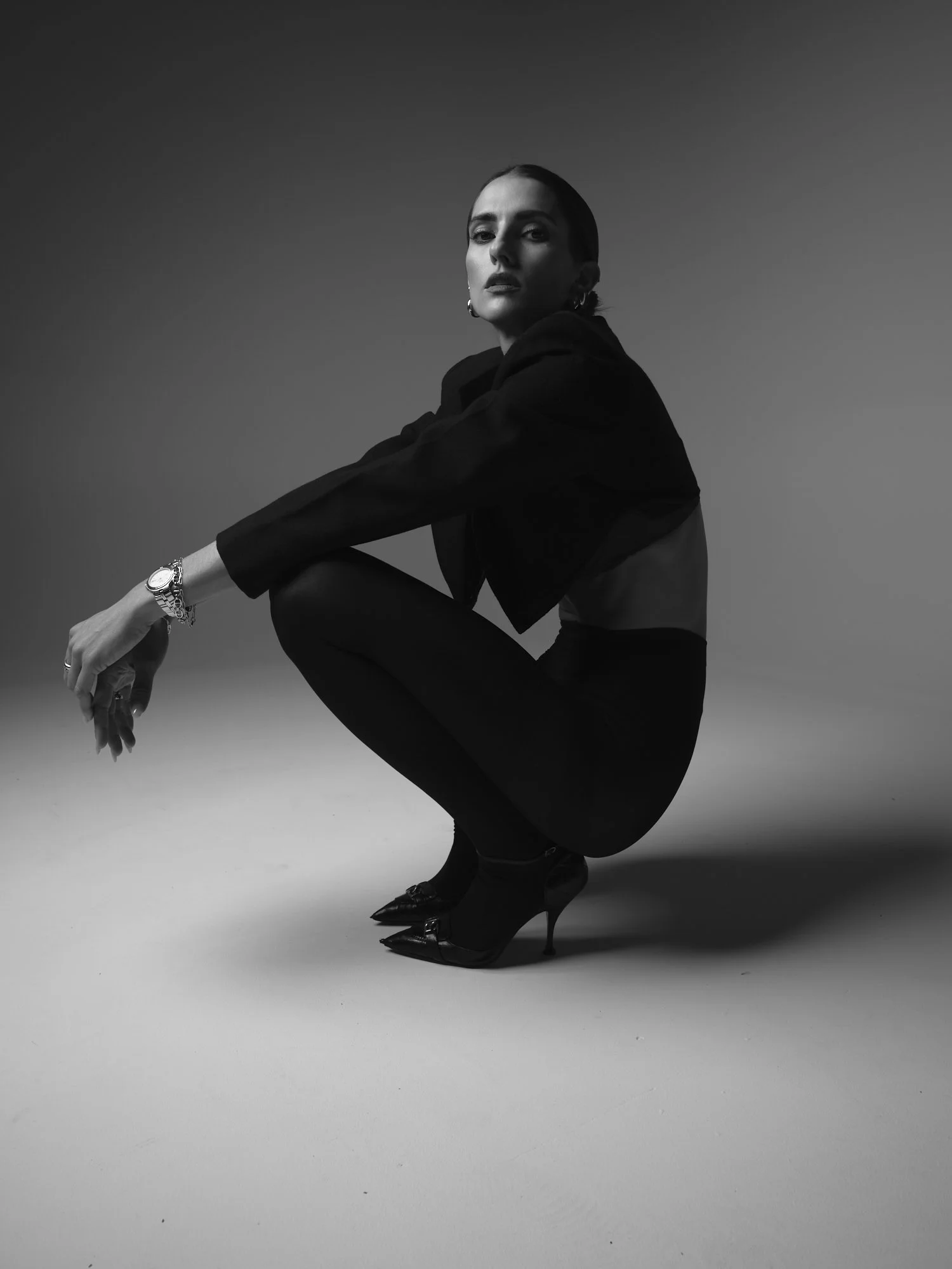 A black and white photo of a woman squatting, wearing high heels, a dark jacket, and jewelry, with a plain gradient background.