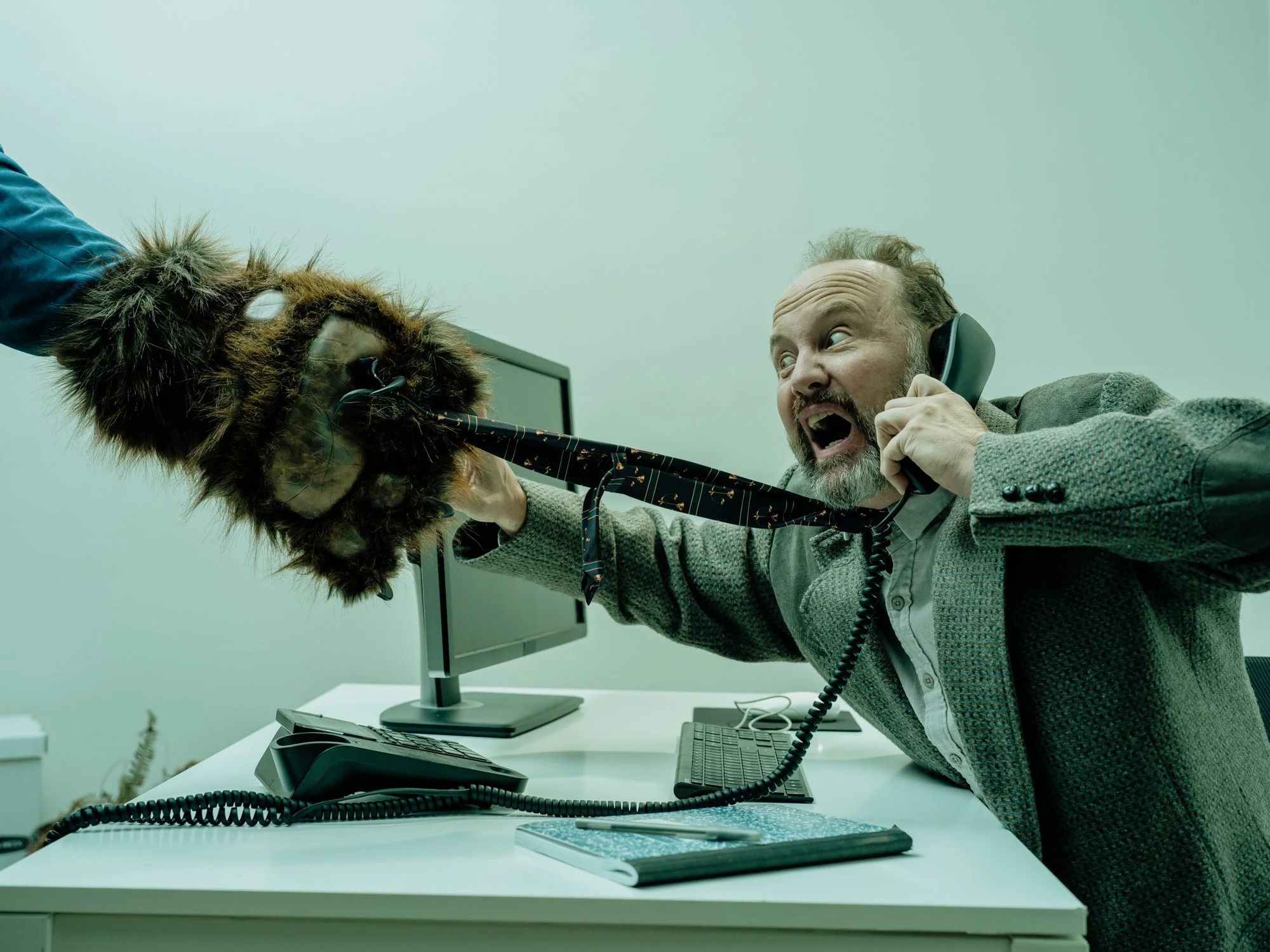 A man in a gray blazer and white shirt is yelling into a black corded phone, holding it with his right hand. He has a beard and mustache, and is sitting at a white desk in an office with a computer monitor, keyboard, and mouse. There are some small d