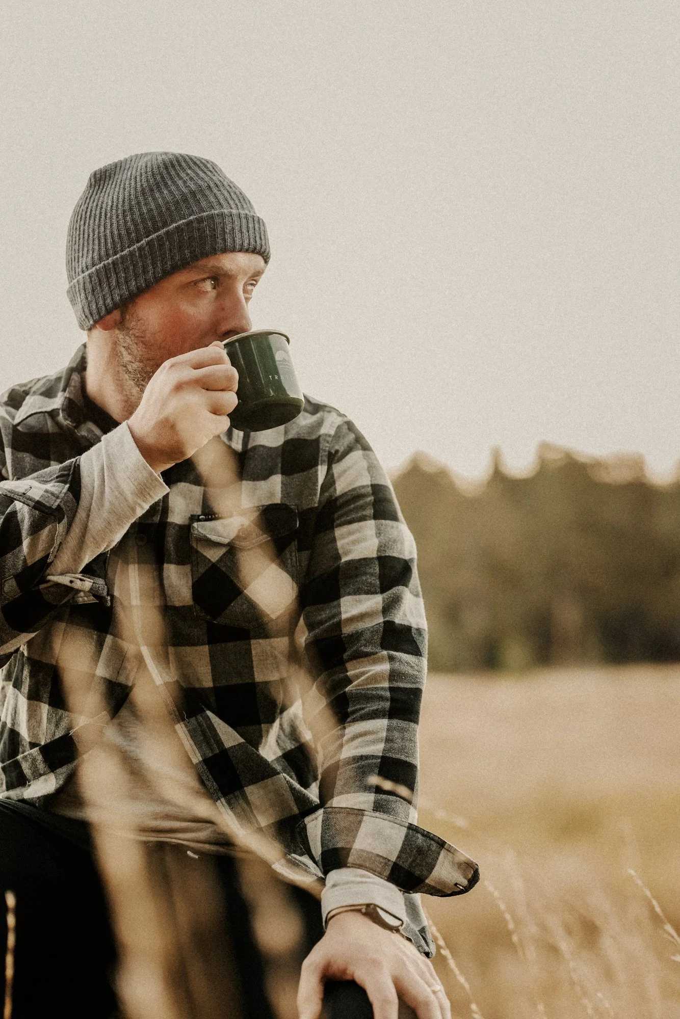 A man wearing a gray beanie and flannel shirt, drinking from a black cup outdoors in a field, with blurred distant trees and a cloudy sky in the background.