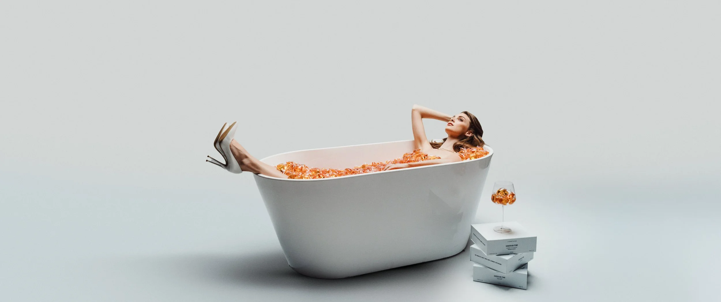 A woman lying in a white bathtub filled with orange petals, wearing high heels, with a glass of orange drink on top of white boxes beside the tub.
