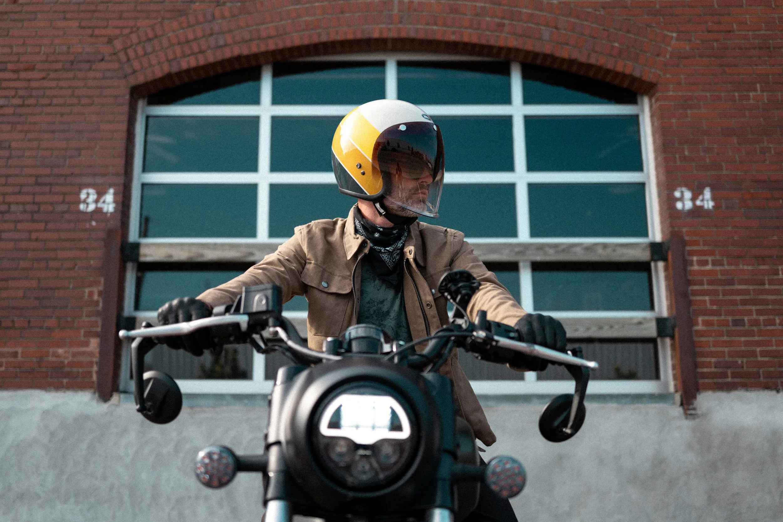 A man wearing a helmet and riding a motorcycle in front of a brick building with the number 34 visible.
