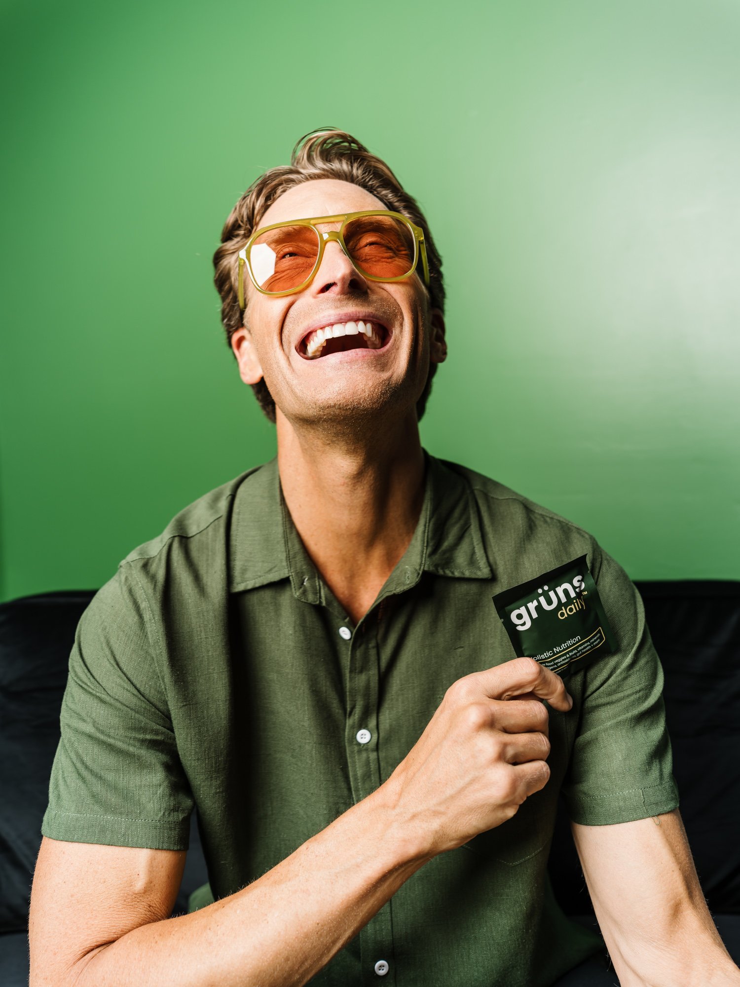 A man with brown hair wearing sunglasses and a green shirt laughs while holding a packet of Grün daily nutritional supplement against a green gradient background.