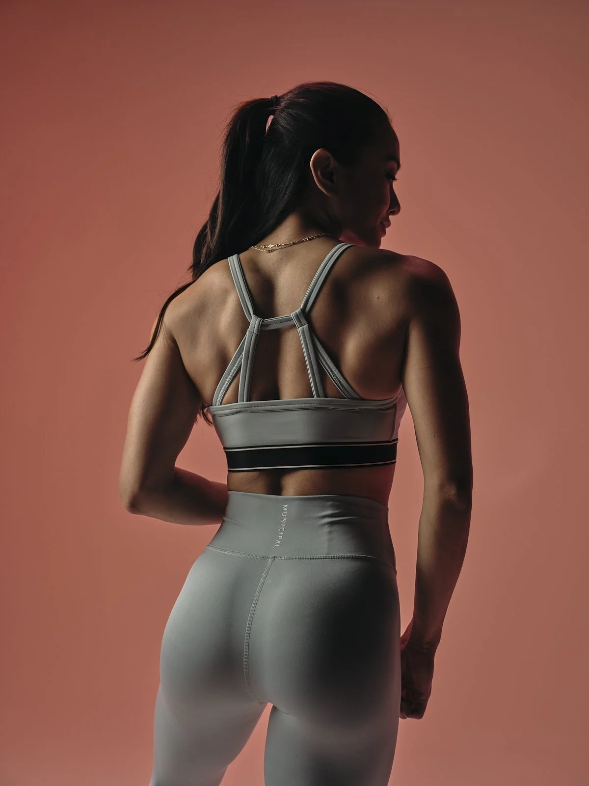 A woman in athletic wear standing against a pink background, turning slightly to her right, showing her back and side profile.