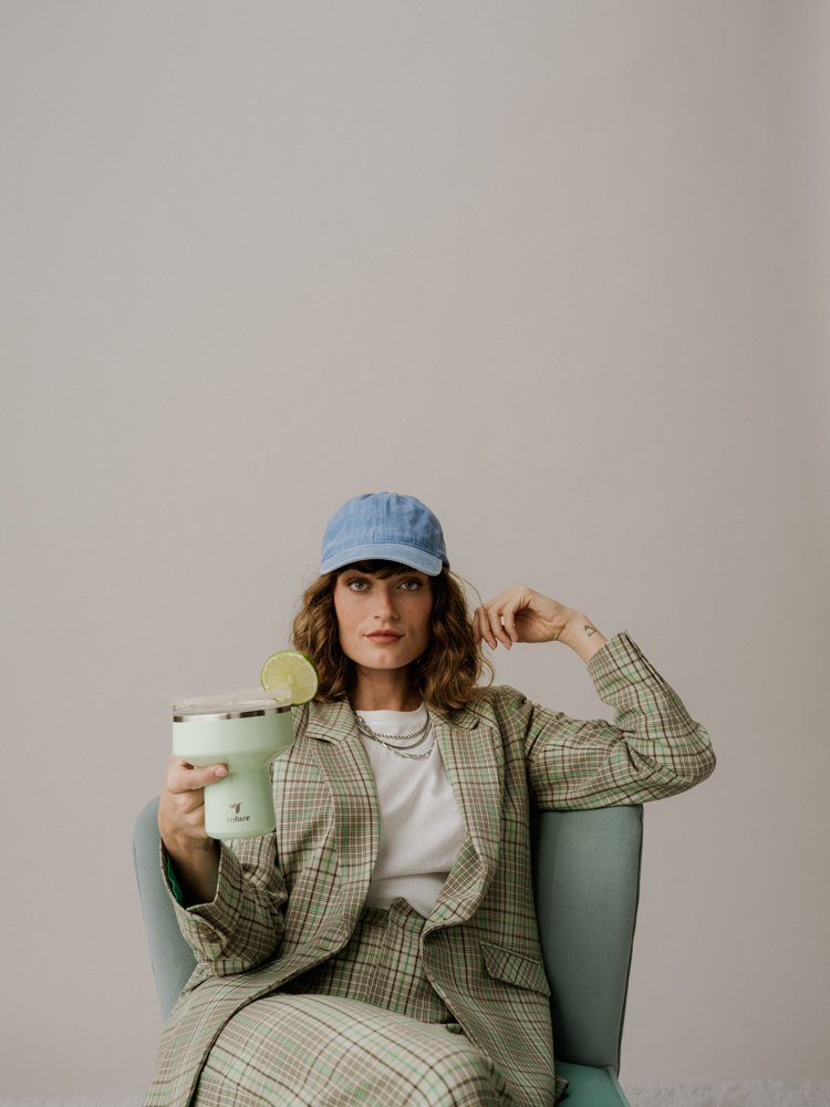 A woman wearing a blue baseball cap, plaid blazer, and white shirt, sitting in a light green chair, holding a margarita glass with a lime wedge.