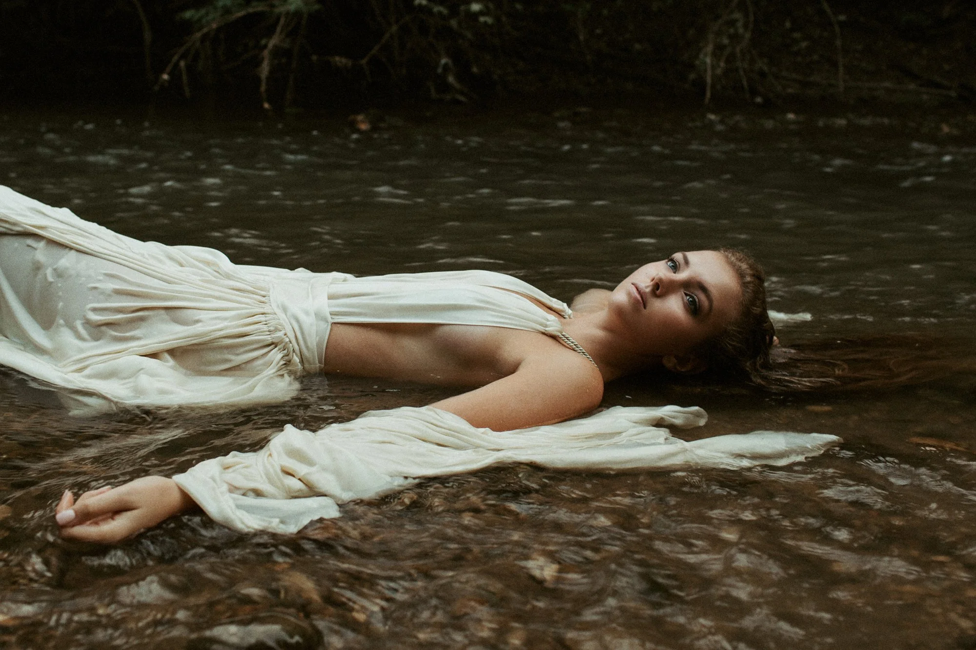 A woman with light skin and curly hair lying in a shallow stream, wearing a cream-colored dress with an intense gaze.