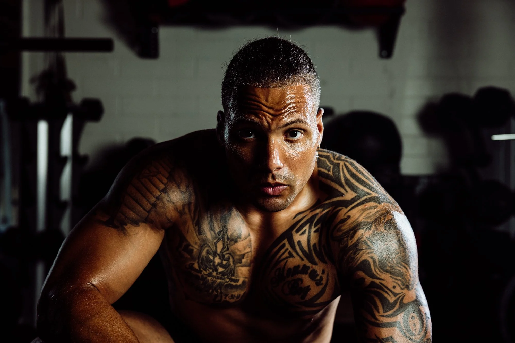A tattooed man in a gym setting, looking intensely at the camera, with gym equipment blurred in the background.