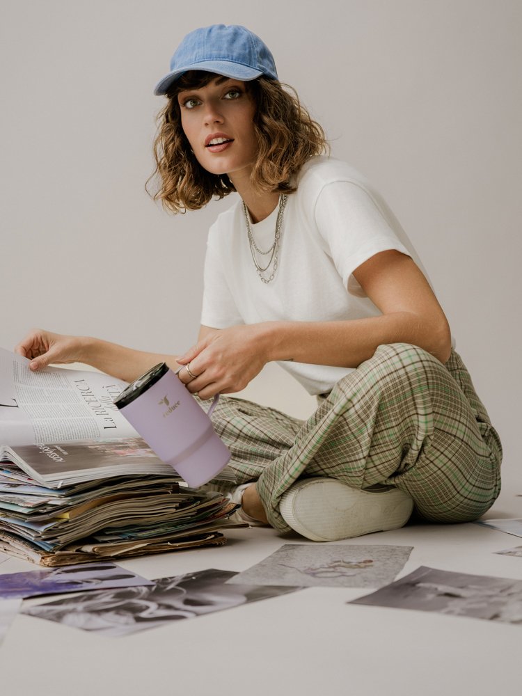 A woman with curly hair wearing a blue cap, white t-shirt, and plaid pants, sitting on the floor surrounded by papers, magazines, and photographs, holding a purple tumbler.