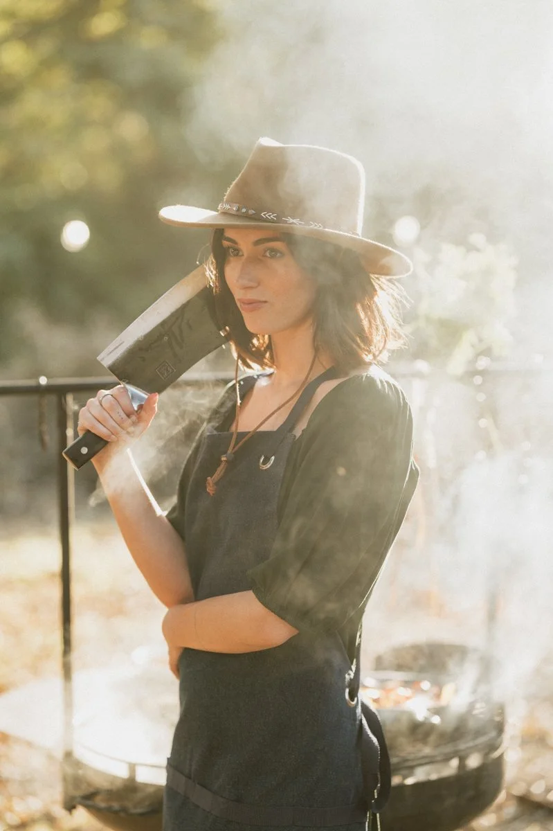 A woman wearing a beige wide-brimmed hat and black overalls, holding a meat cleaver, standing outdoors near a grill with smoke.