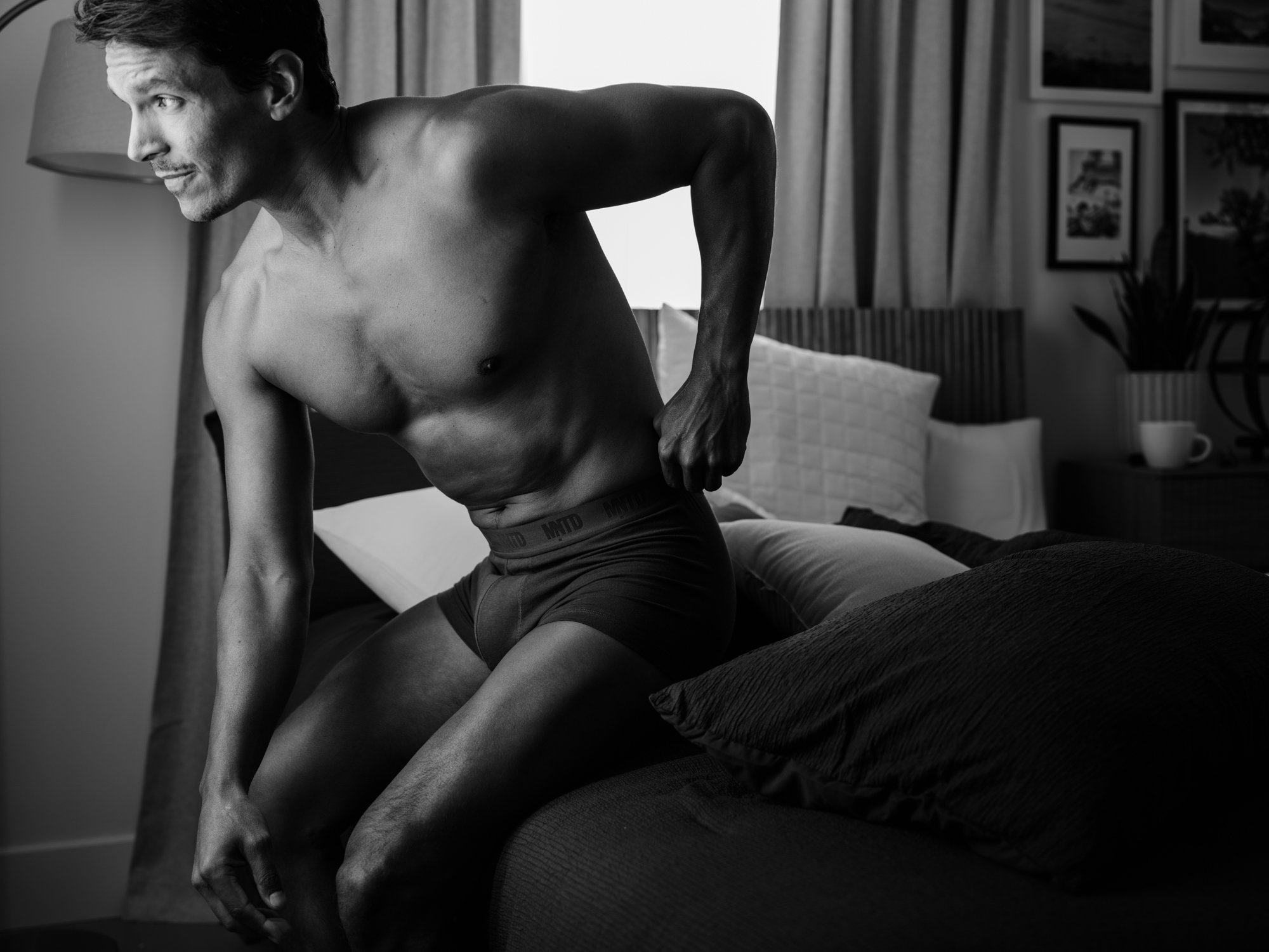 A shirtless man sitting on a bed, looking to the left, in a bedroom with framed pictures and a plant in the background.