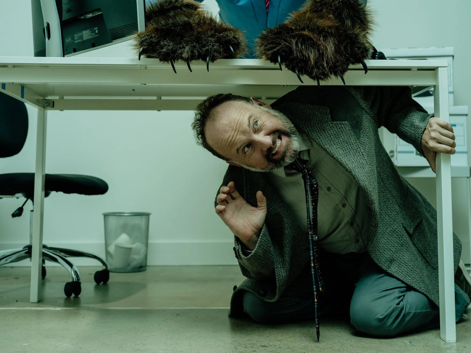 A man is crouching under a white desk, looking up with a surprised or playful expression. The desk has animal claws and fur on top, resembling a creature's paws. Behind him are office chairs, a trash can, and a computer monitor.