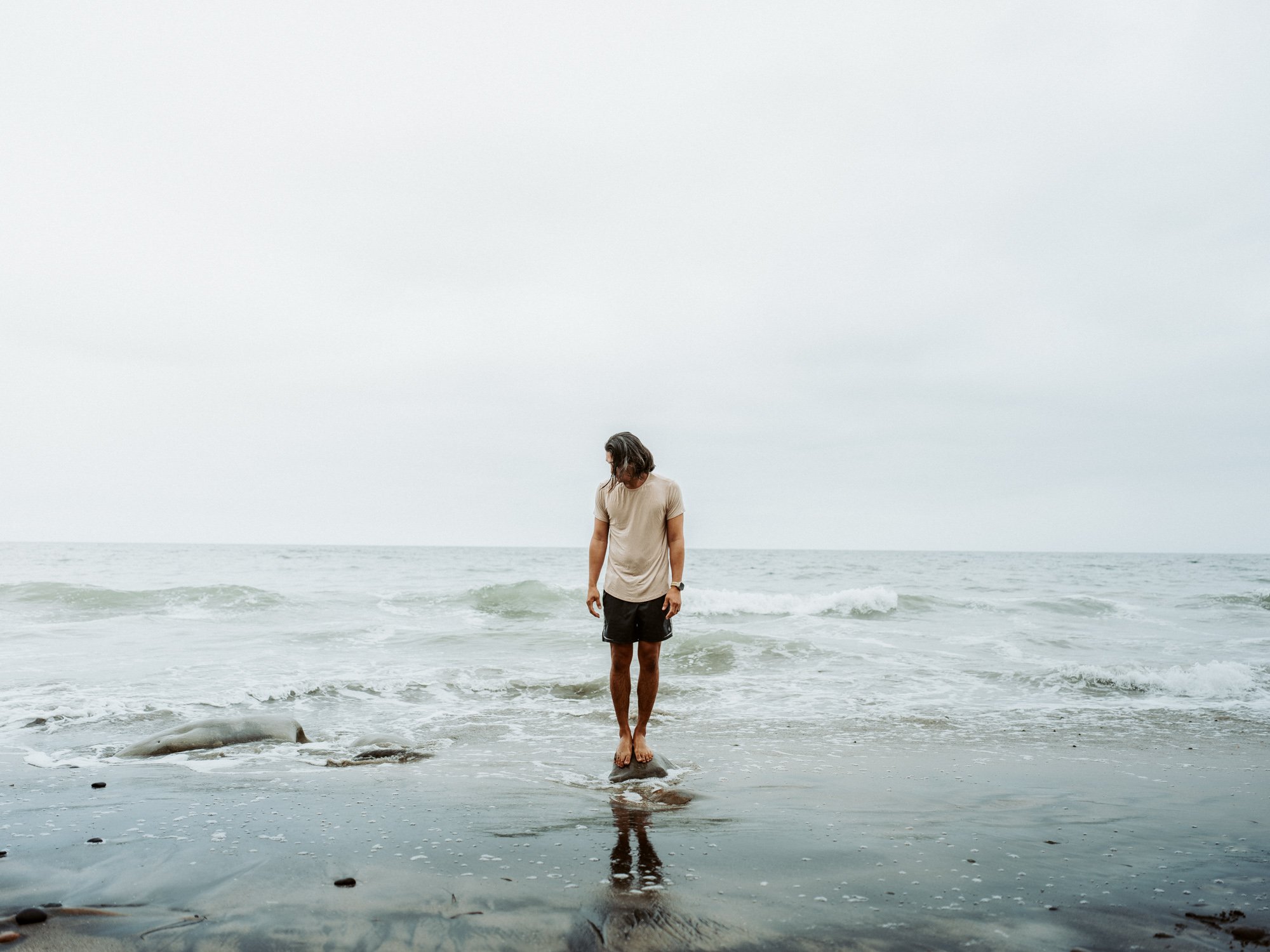 A person with long hair on the beach, standing on rocks at the shoreline, looking down at the water with a calm expression, during an overcast day.