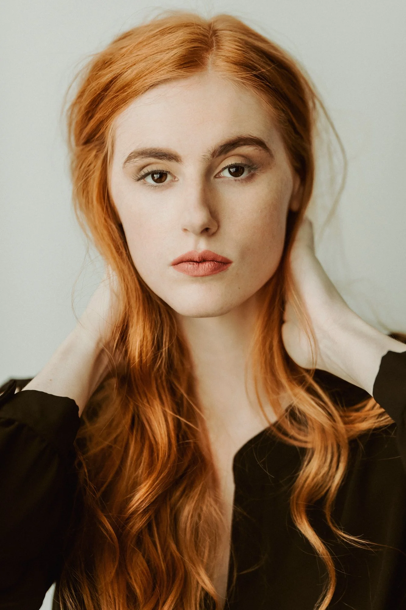 A woman with long wavy red hair, light skin, green eyes, and wearing a black top, looking directly at the camera with her hands behind her neck.