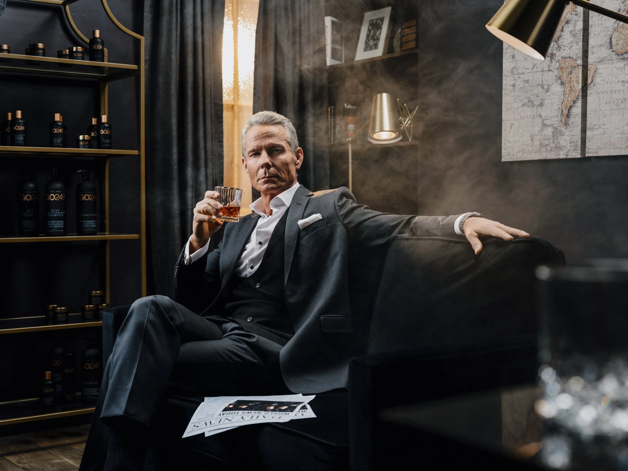 A man in a gray suit sitting on a black leather sofa in a modern room, holding a glass of whiskey, with bottles of GQ24 hair products on the shelves behind him, a map on the wall, and a paper on the table.