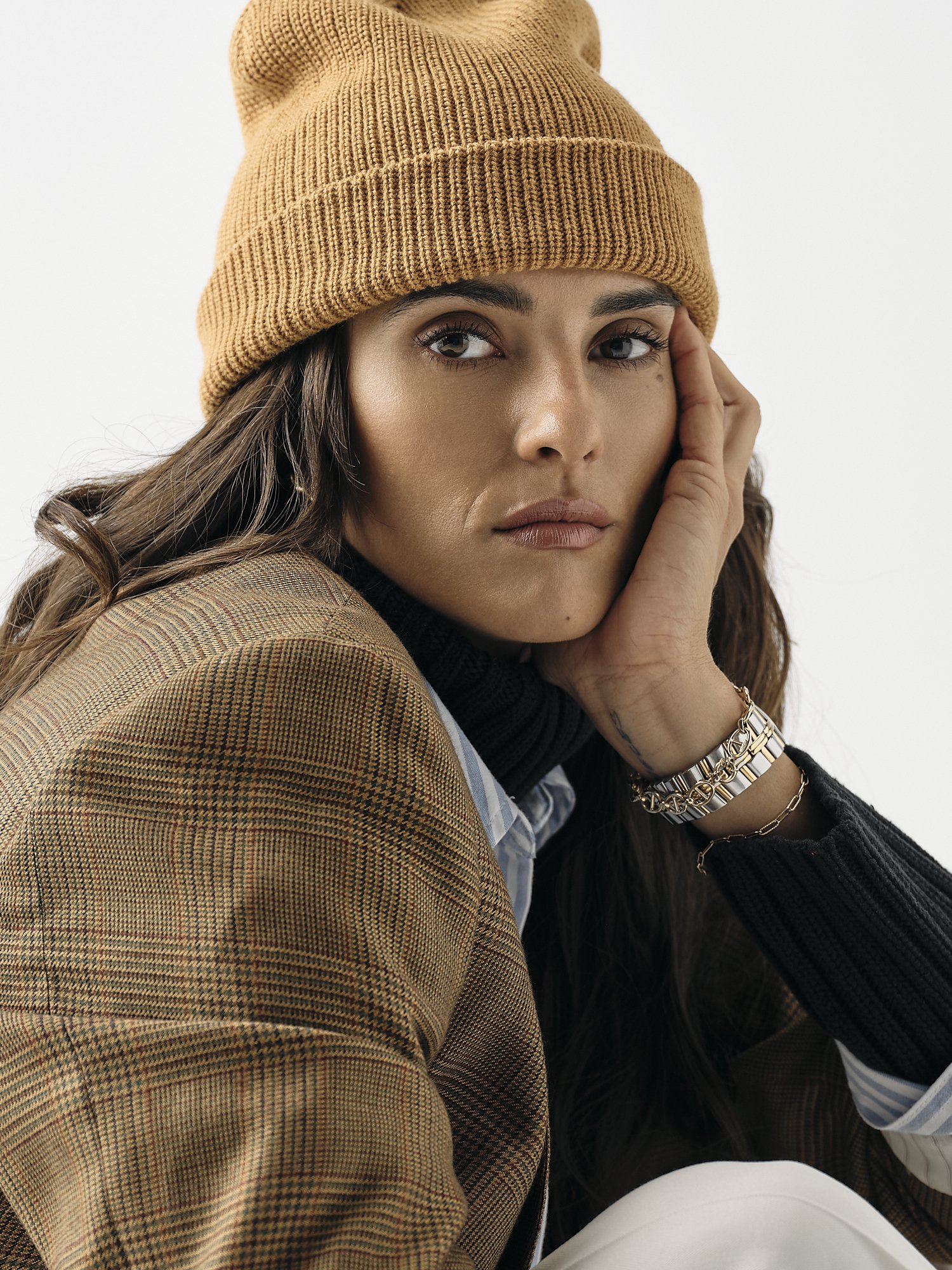 Close-up of a woman wearing a beige knit beanie, a plaid blazer, and jewelry, with her hand resting near her ear.