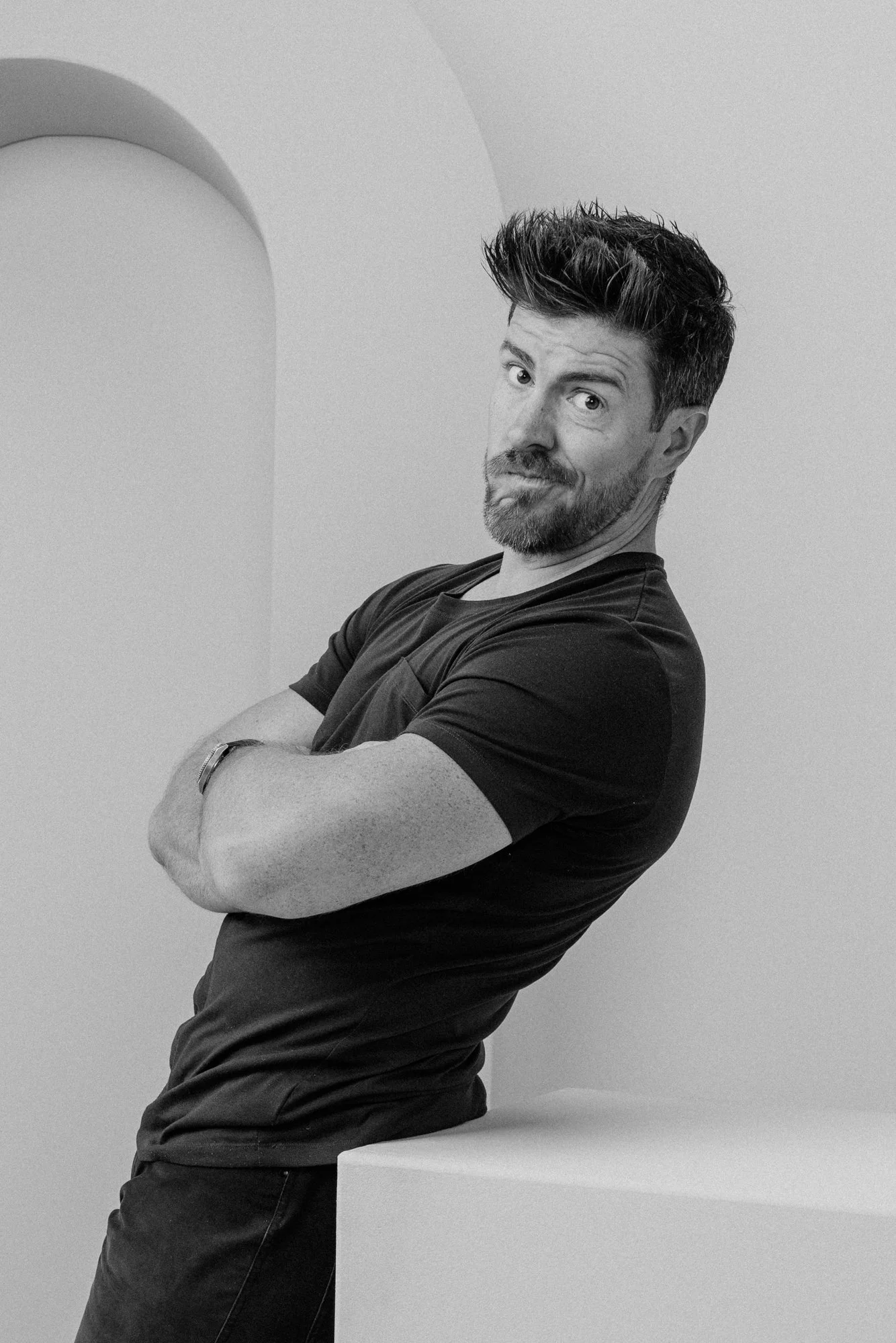 A man with dark, styled hair and a beard, wearing a black T-shirt, sitting on a white surface with his arms crossed, looking at the camera with a playful expression, in a minimalist room with white walls and an arch detail.