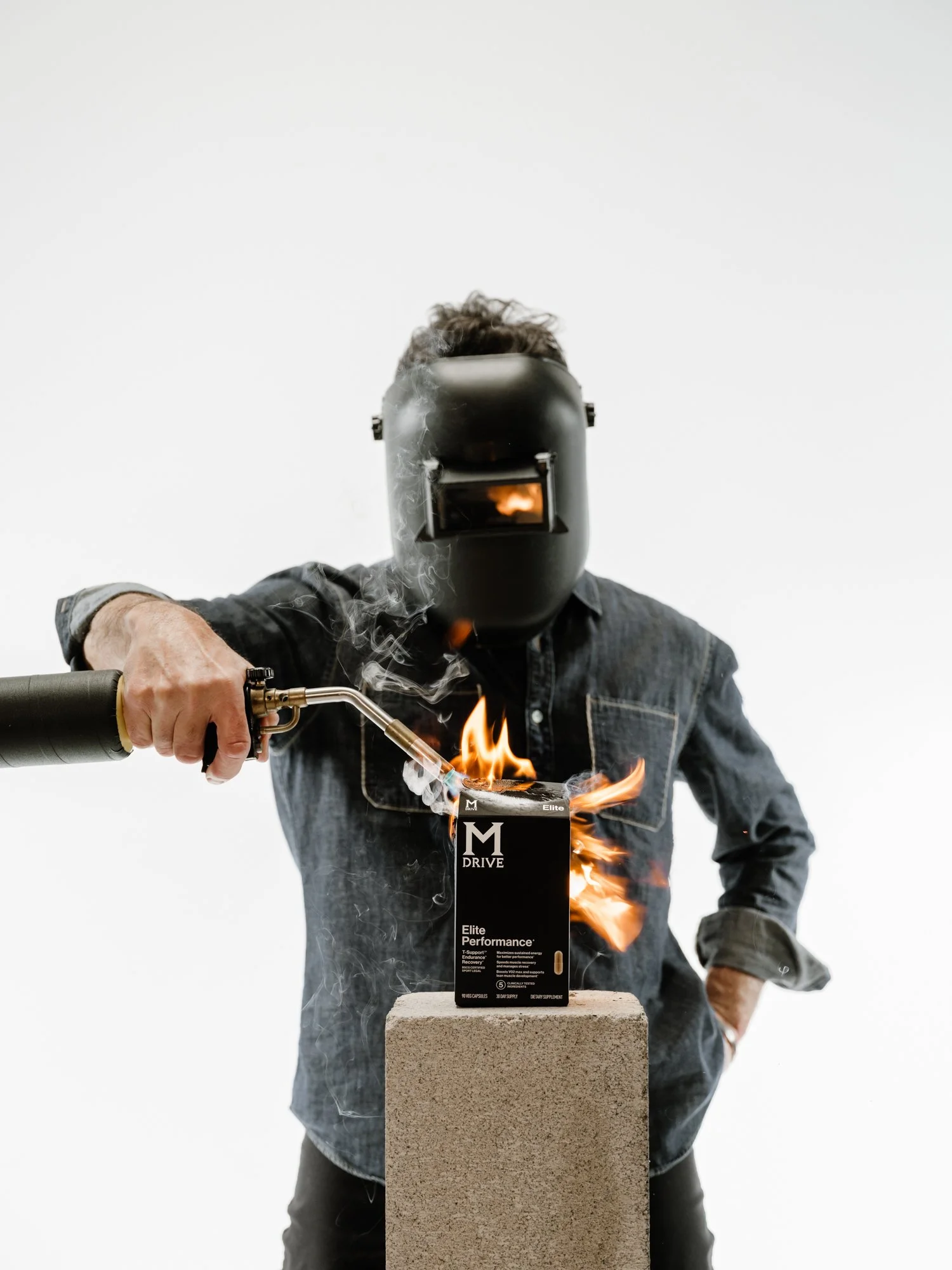 A person wearing a welding helmet, holding a blowtorch, and igniting a box of dietary supplements on a concrete block.