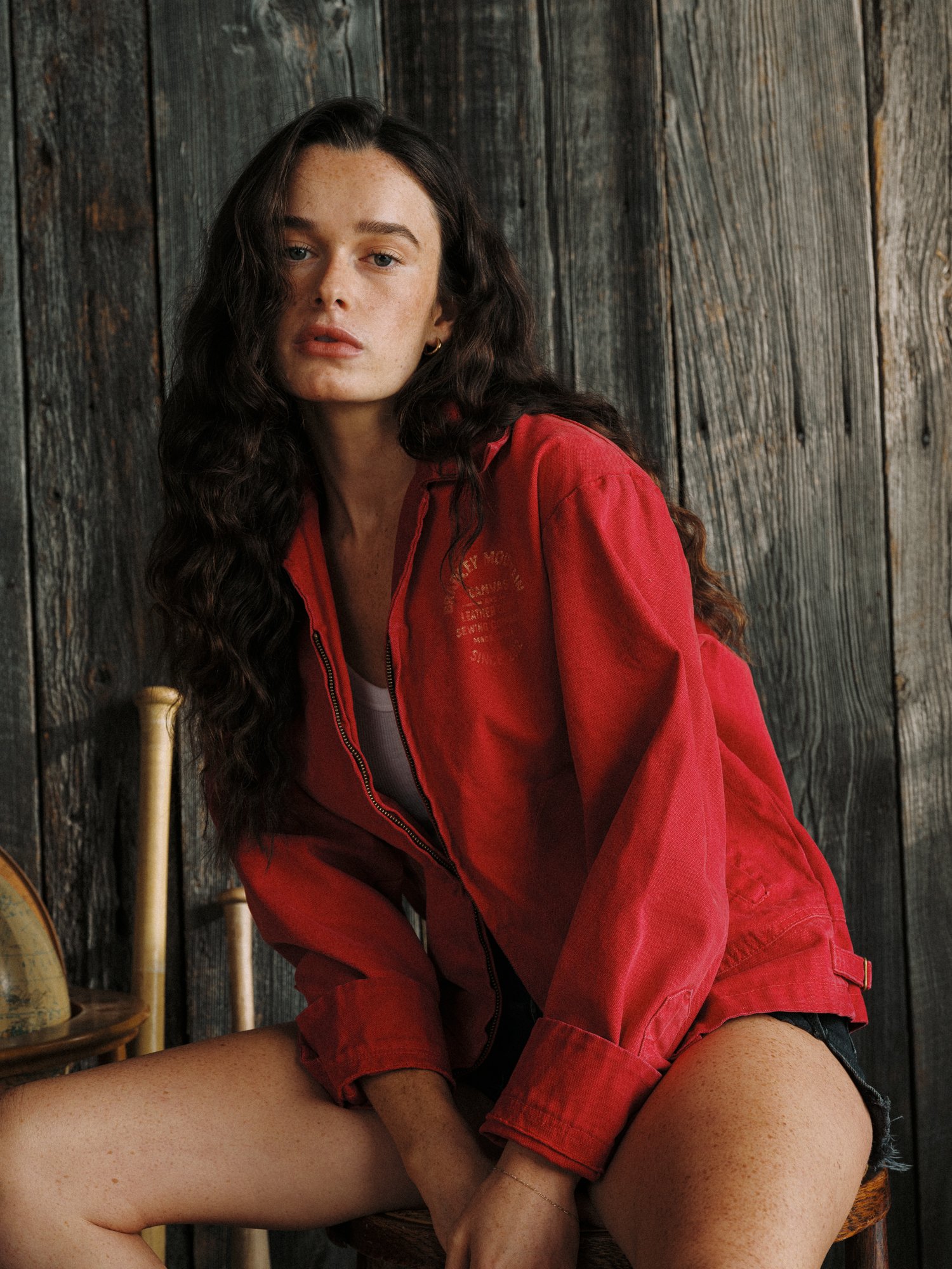 A young woman with long curly dark hair, wearing a red jacket, sitting against a rustic wooden wall, with legs crossed and looking at the camera.
